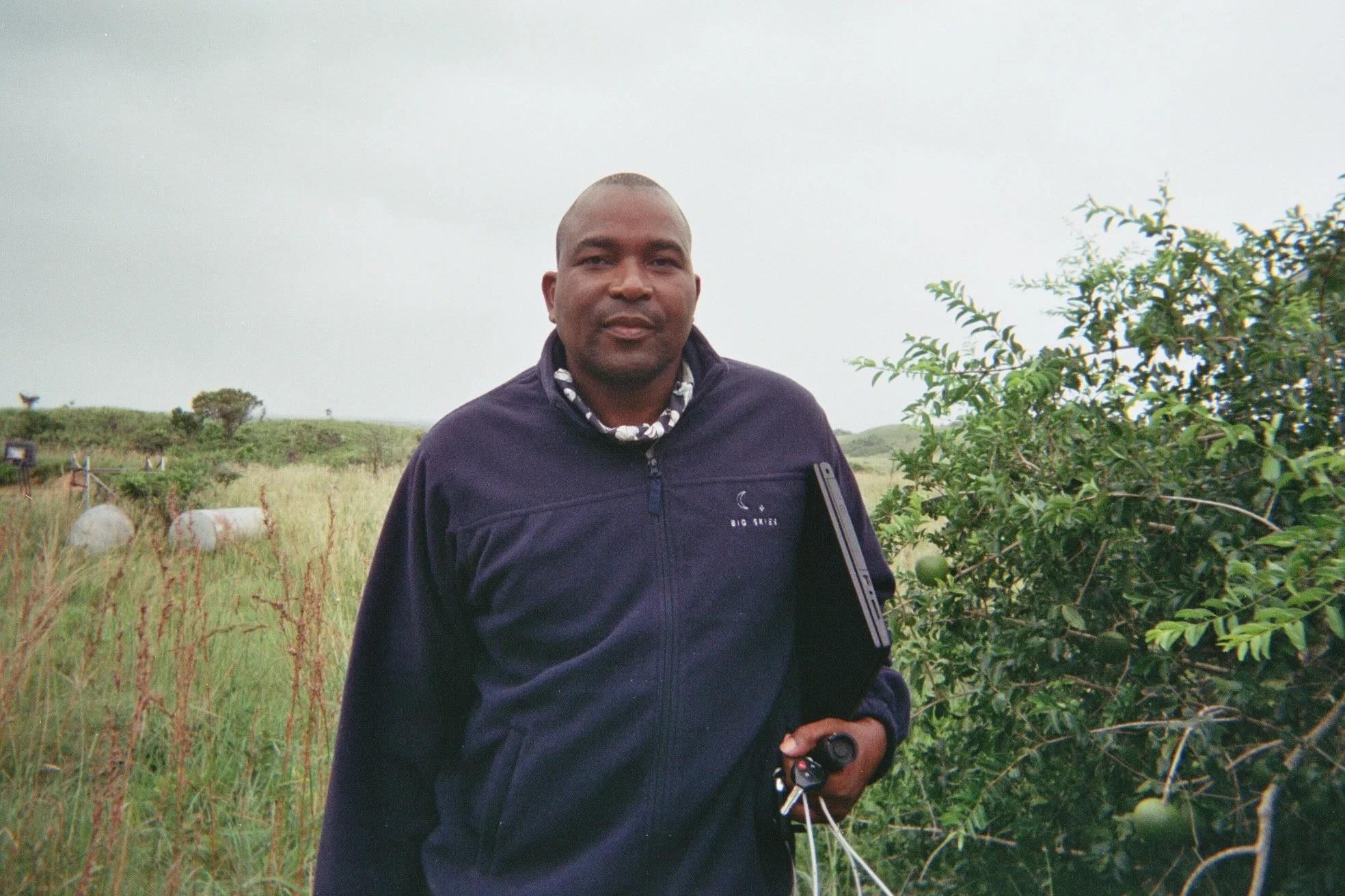 A man standing outdoors in a grassy field with bushes and a cloudy sky in the background, holding a drone controller in his hand.