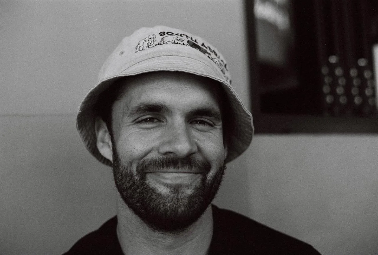 A smiling man with a beard wearing a bucket hat, indoors with a blurred background.