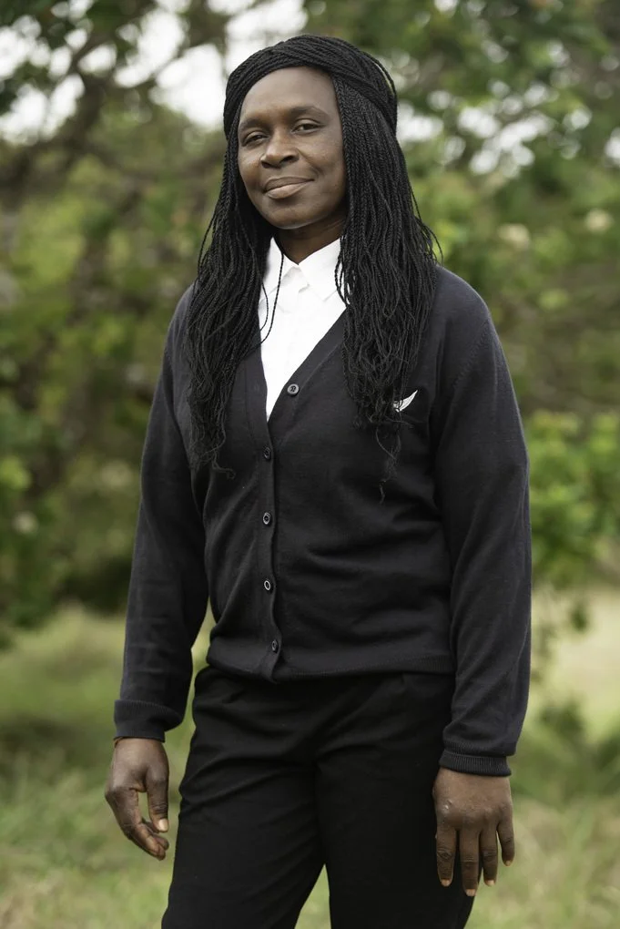 A person wearing a black cardigan and white shirt stands outdoors with greenery in the background.