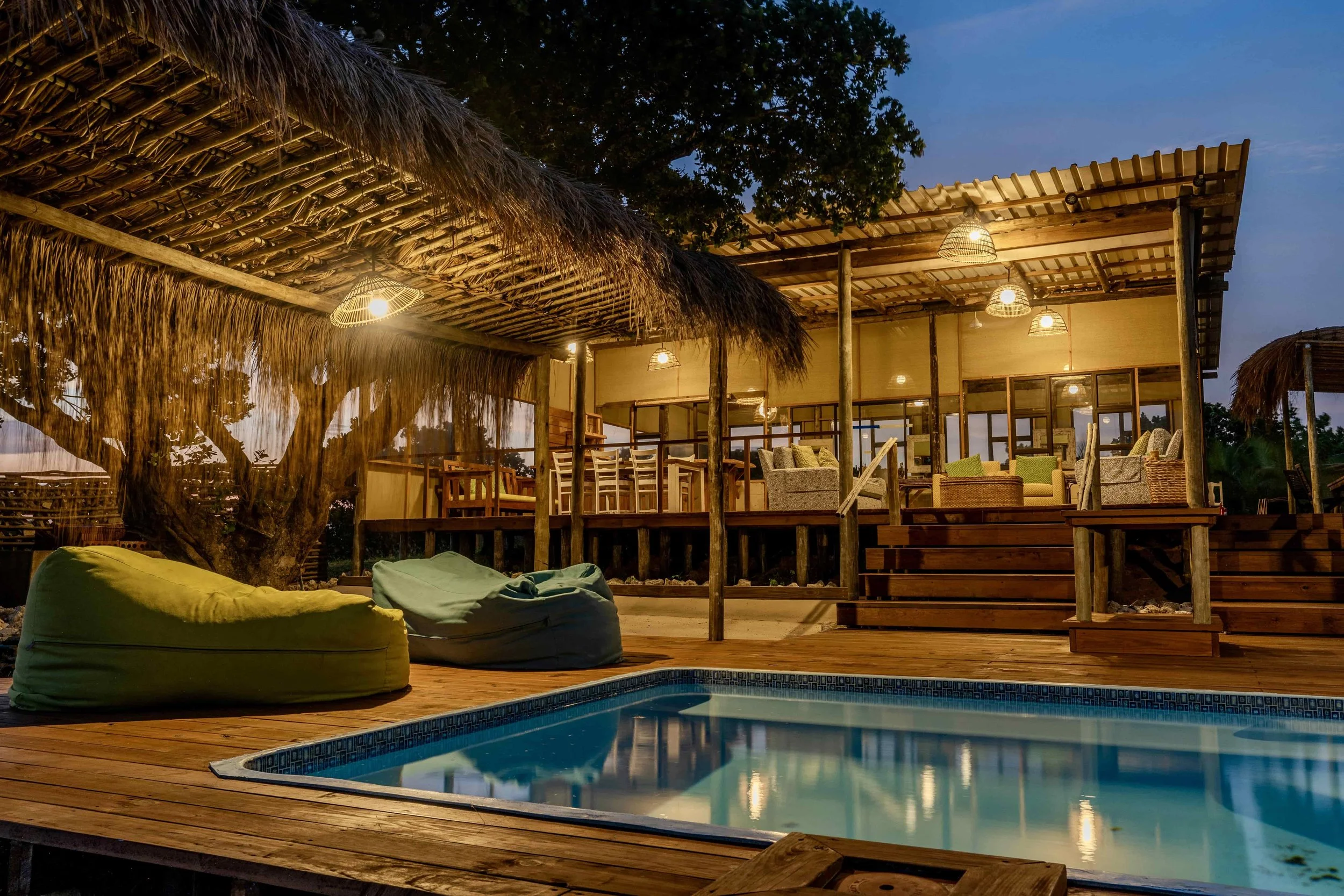Rustic outdoor lounge area with wooden deck, swimming pool, wicker furniture, bean bags, and thatched roof at dusk.