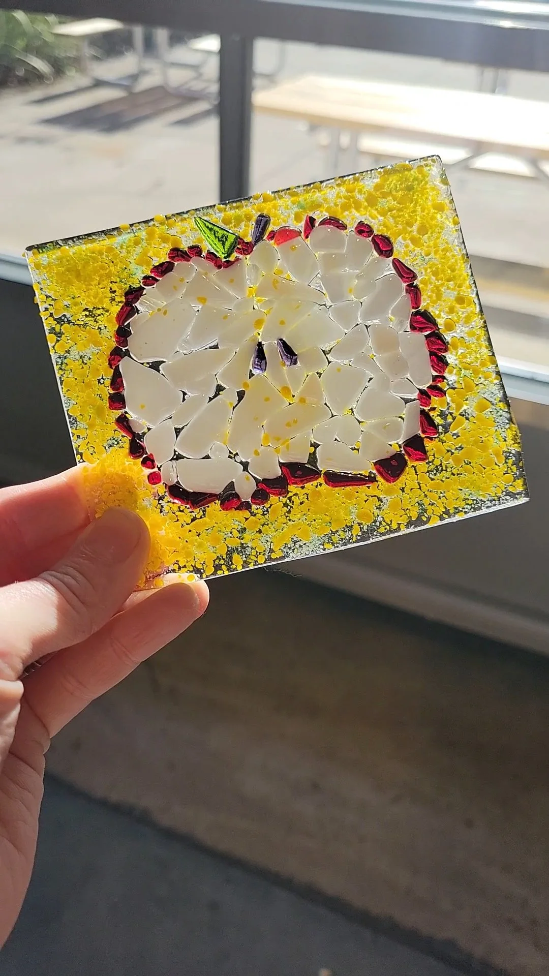 Pictured is a cross section of an apple designed in glass on a clear black canvas sprinkled with yellow frit, and decorated with red, white, and green glass bits to create the image of an apple.