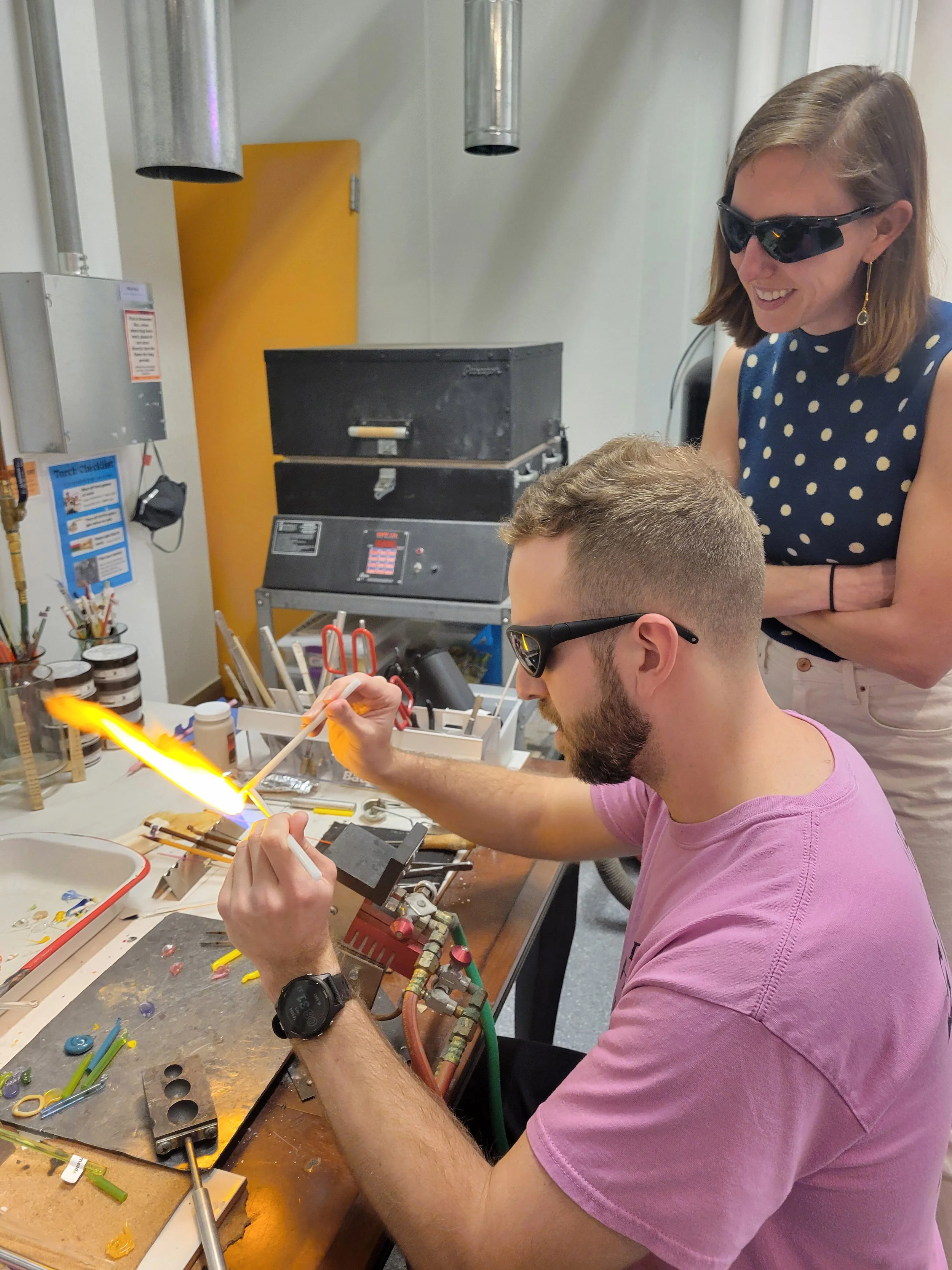 A class participant sits at a table top torch producing a flame.  He is manipulating two glass rods in the flame as another participant looks on.