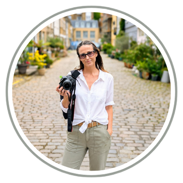 Woman with glasses holding a camera, standing on a cobblestone street with potted plants and buildings in the background.