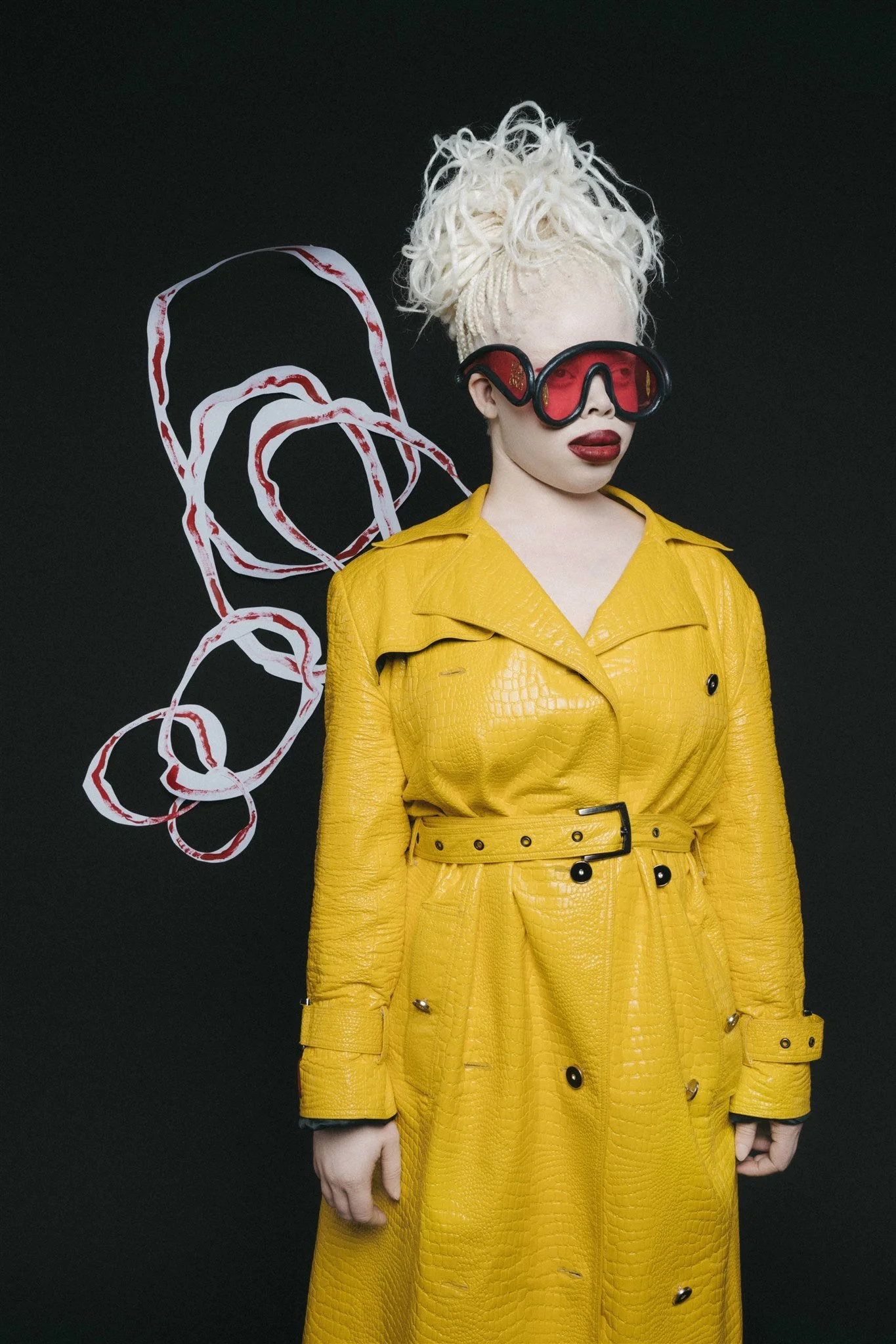 Personne portant un manteau jaune, lunettes de protection rouges, fond noir décoré de cercles rouges et blancs.