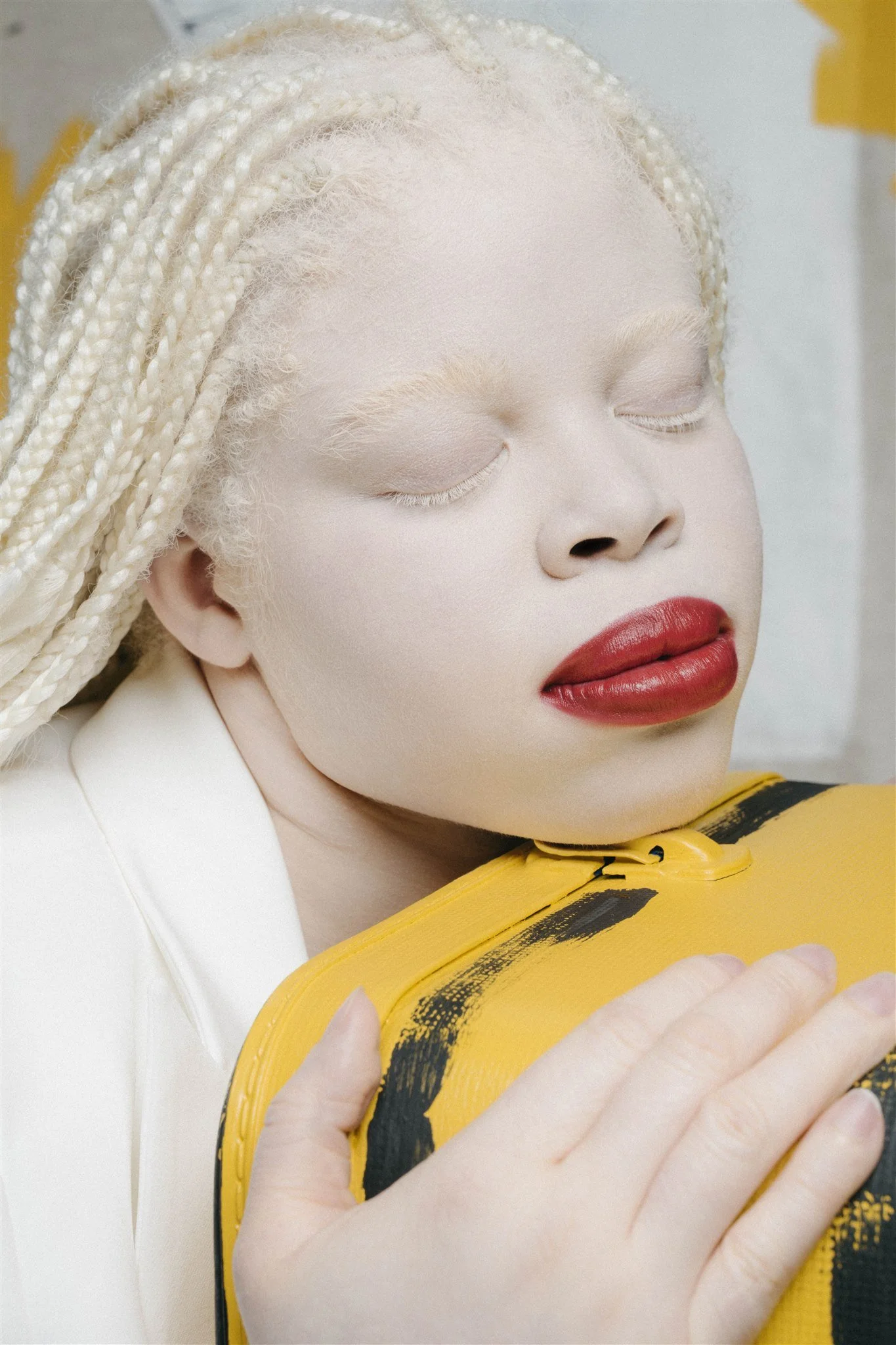 Une personne aux yeux fermés avec des cheveux tressés blonds, maquillée avec du rouge à lèvres rouge, tenant un sac jaune avec des motifs noirs.