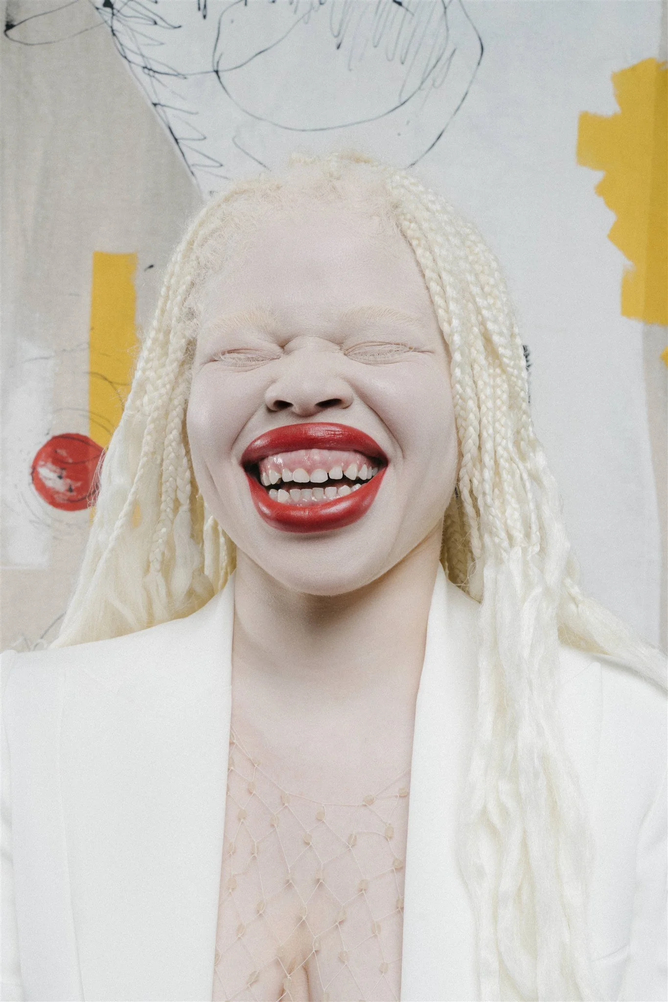 Personne souriante aux cheveux tressés blonds, vêtu d'un vêtement blanc devant un fond artistique aux couleurs vives.