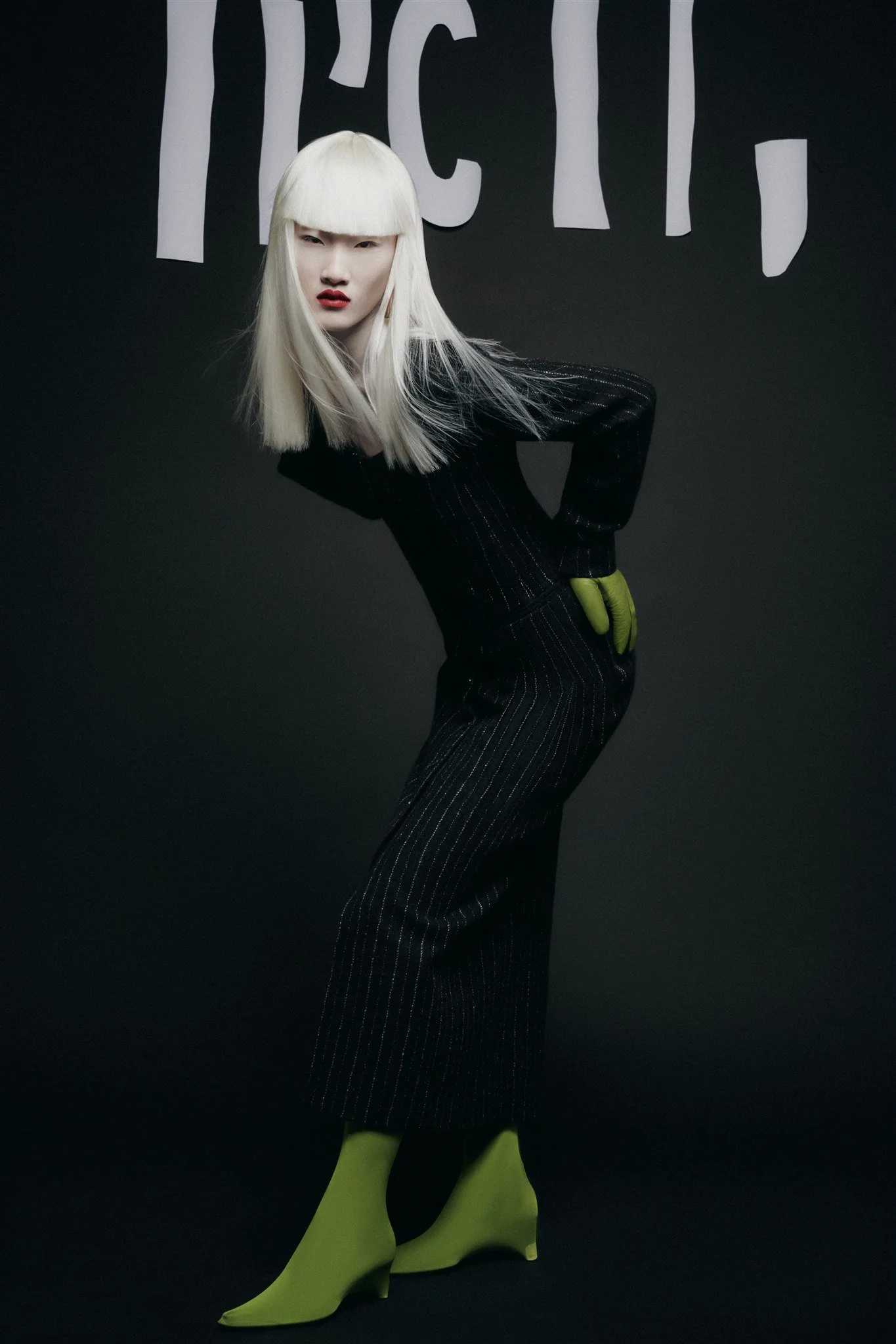 Personne en robe noire à rayures, gants verts, talons verts, cheveux blonds, devant un fond sombre avec des lettres blanches.