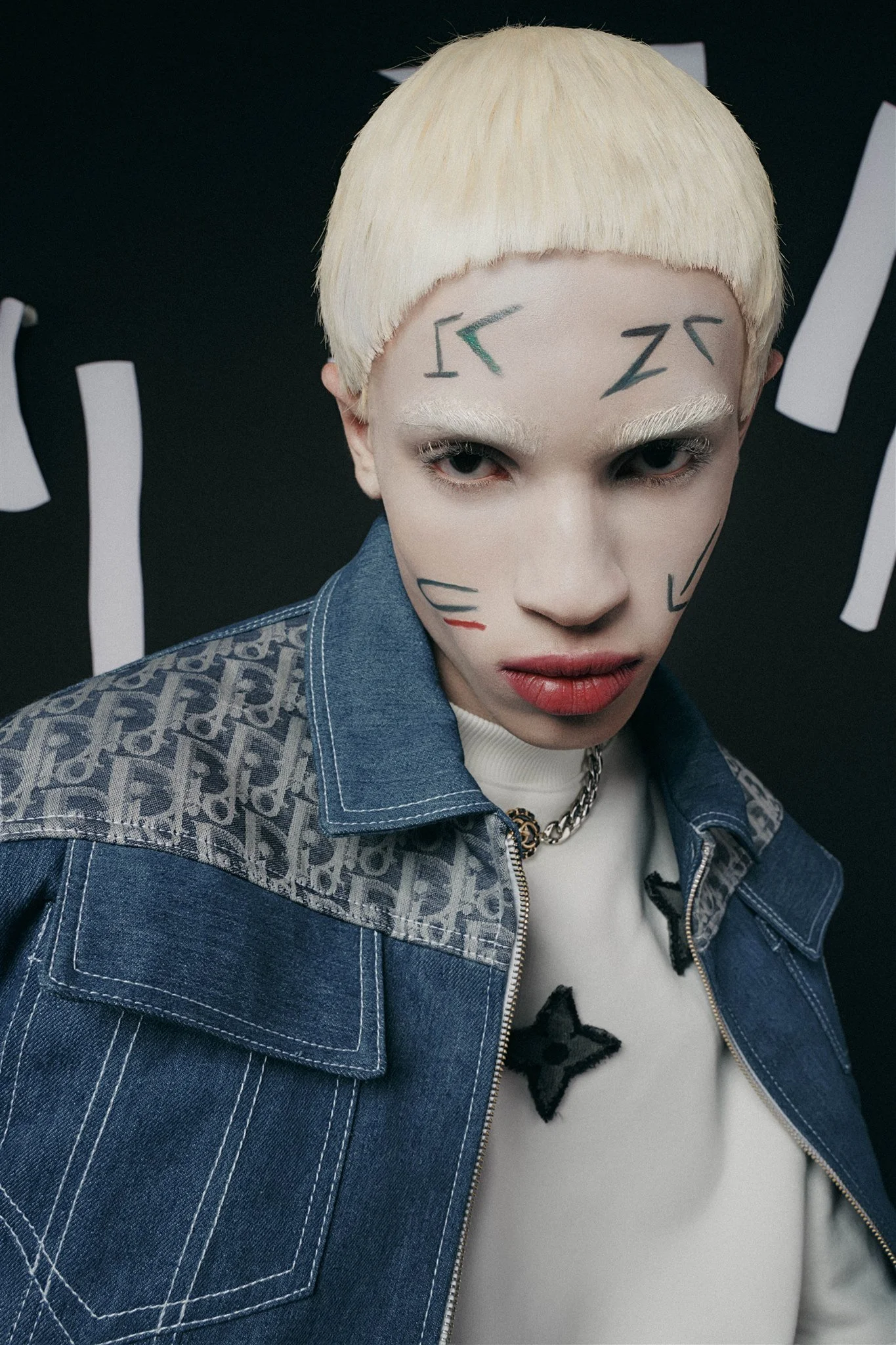 Personne avec des cheveux blonds courts, maquillage graphique sur le visage, portant une veste en jean avec motif et un pull blanc.