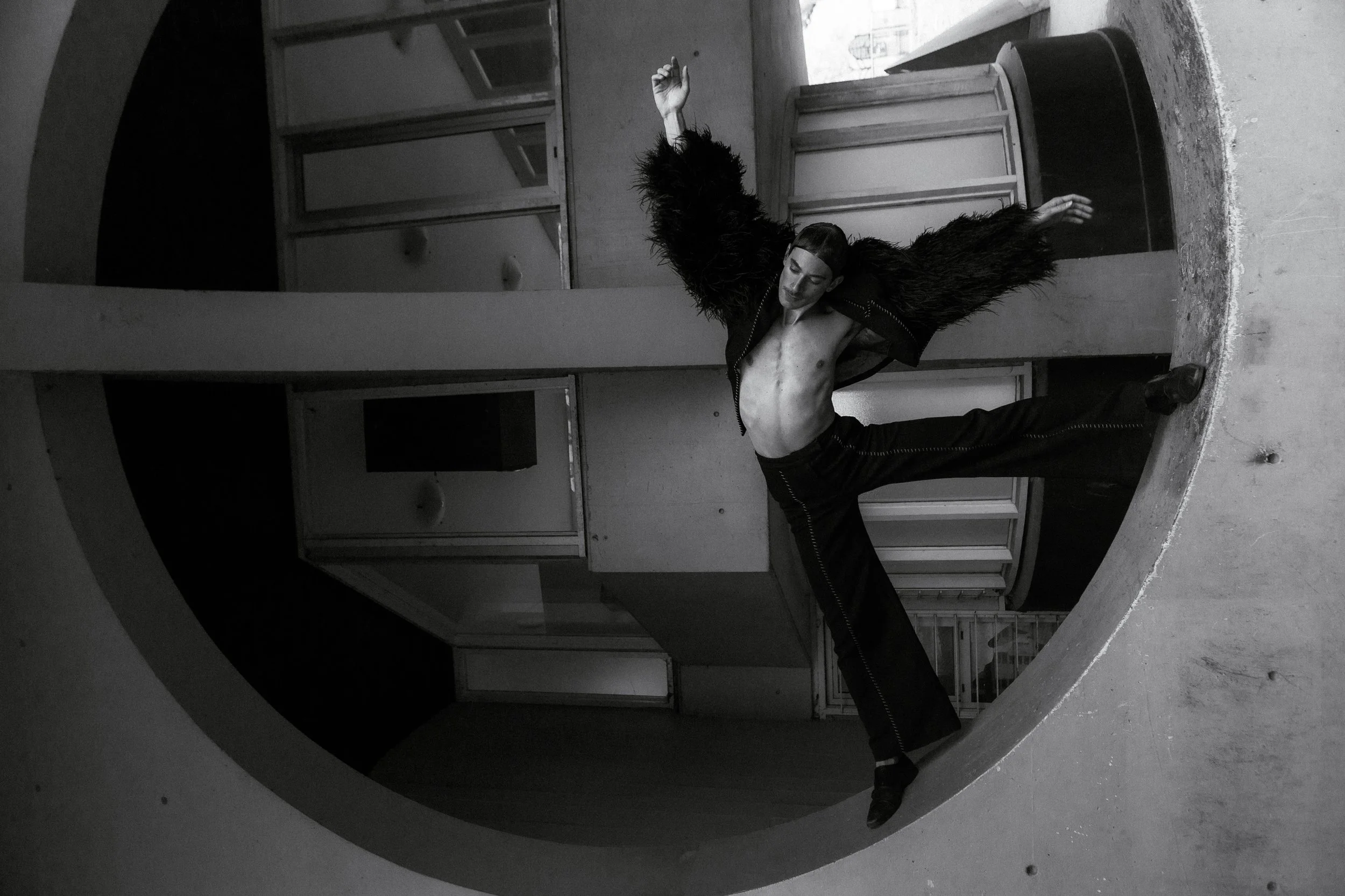 Un danseur en tenue noire effectue une pose acrobatique dans un environnement architectural circulaire en noir et blanc.