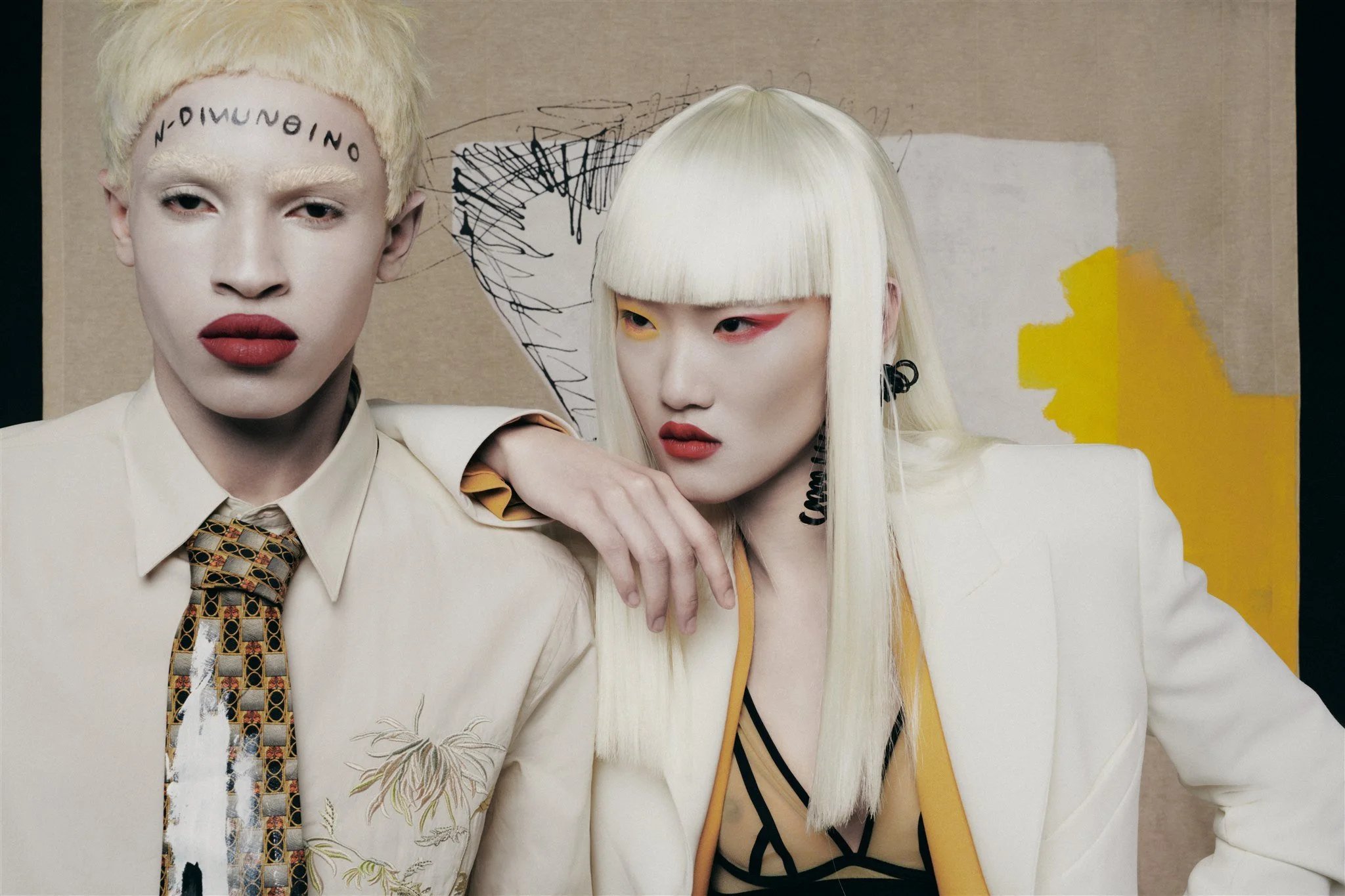 Deux mannequins portant des tenues élégantes, avec du maquillage artistique, posant devant un fond beige et jaune.