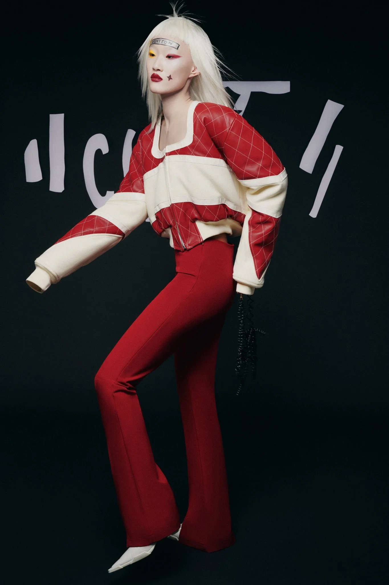 Une personne aux cheveux blonds portant une veste rouge et beige et un pantalon rouge, posant devant un fond noir avec des formes blanches. Maquillage artistique avec un autocollant sur le front.
