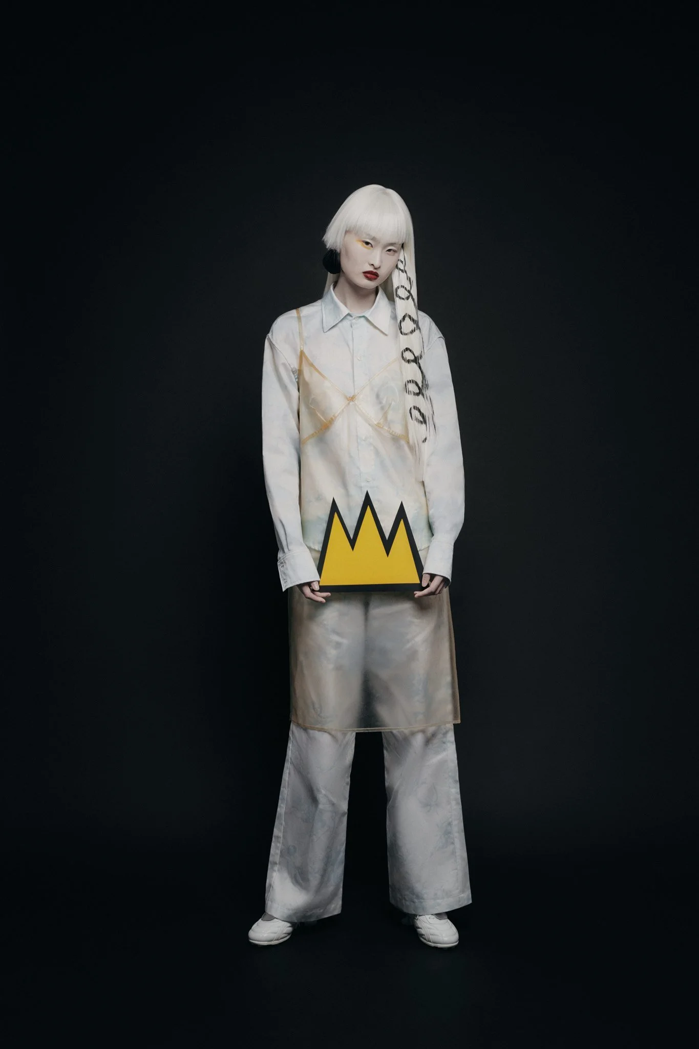 Personne aux cheveux blancs avec un vêtement transparent, tenant un objet jaune en forme de couronne sur fond sombre.
