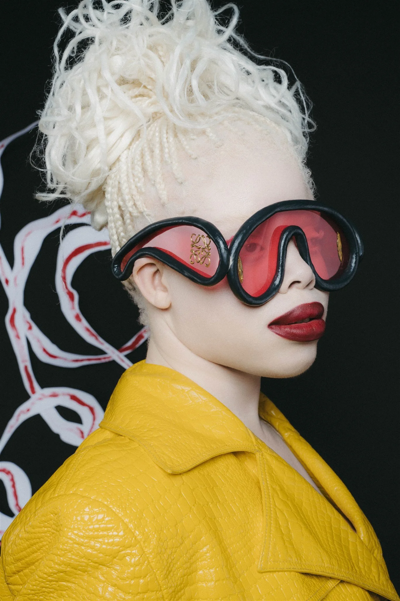 Personne avec cheveux blonds pâles, veste jaune texturée, lunettes de soleil rouges, fond sombre avec motifs blancs et rouges.