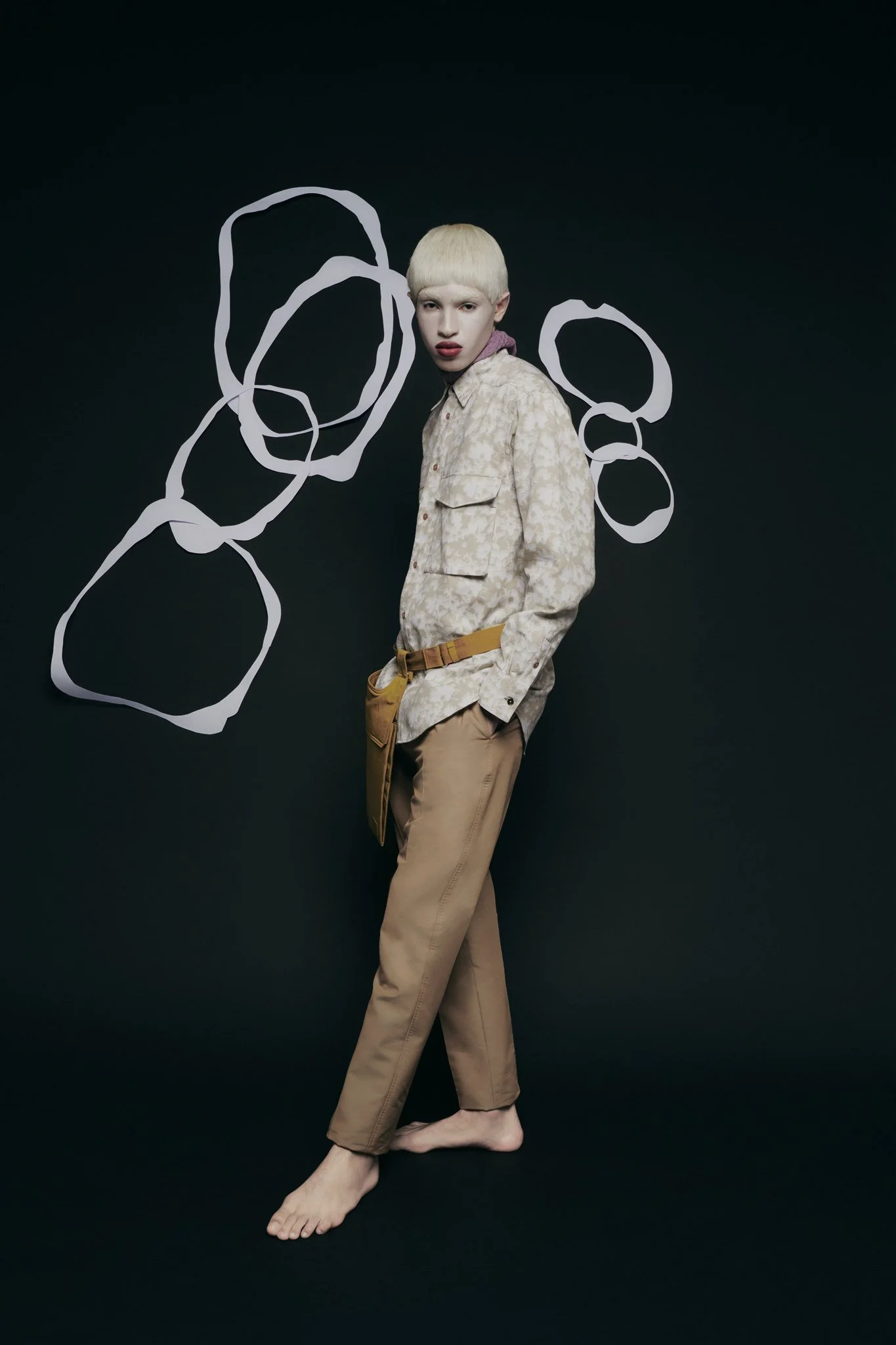 Personne en chemise à motifs avec pantalon beige, pieds nus, posant devant fond sombre avec cercles blancs dessinés.