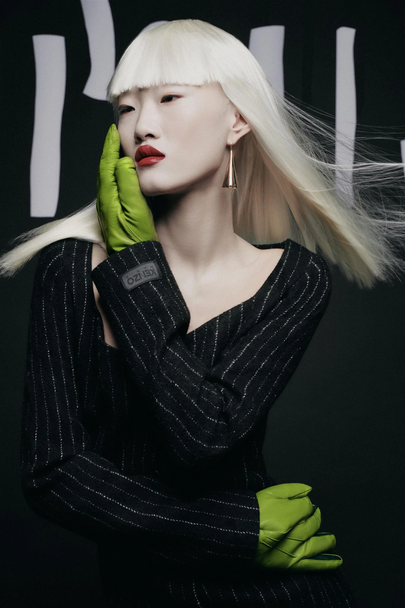 Une personne avec des gants verts, cheveux blonds et rouges à lèvres, portant un pull noir rayé.