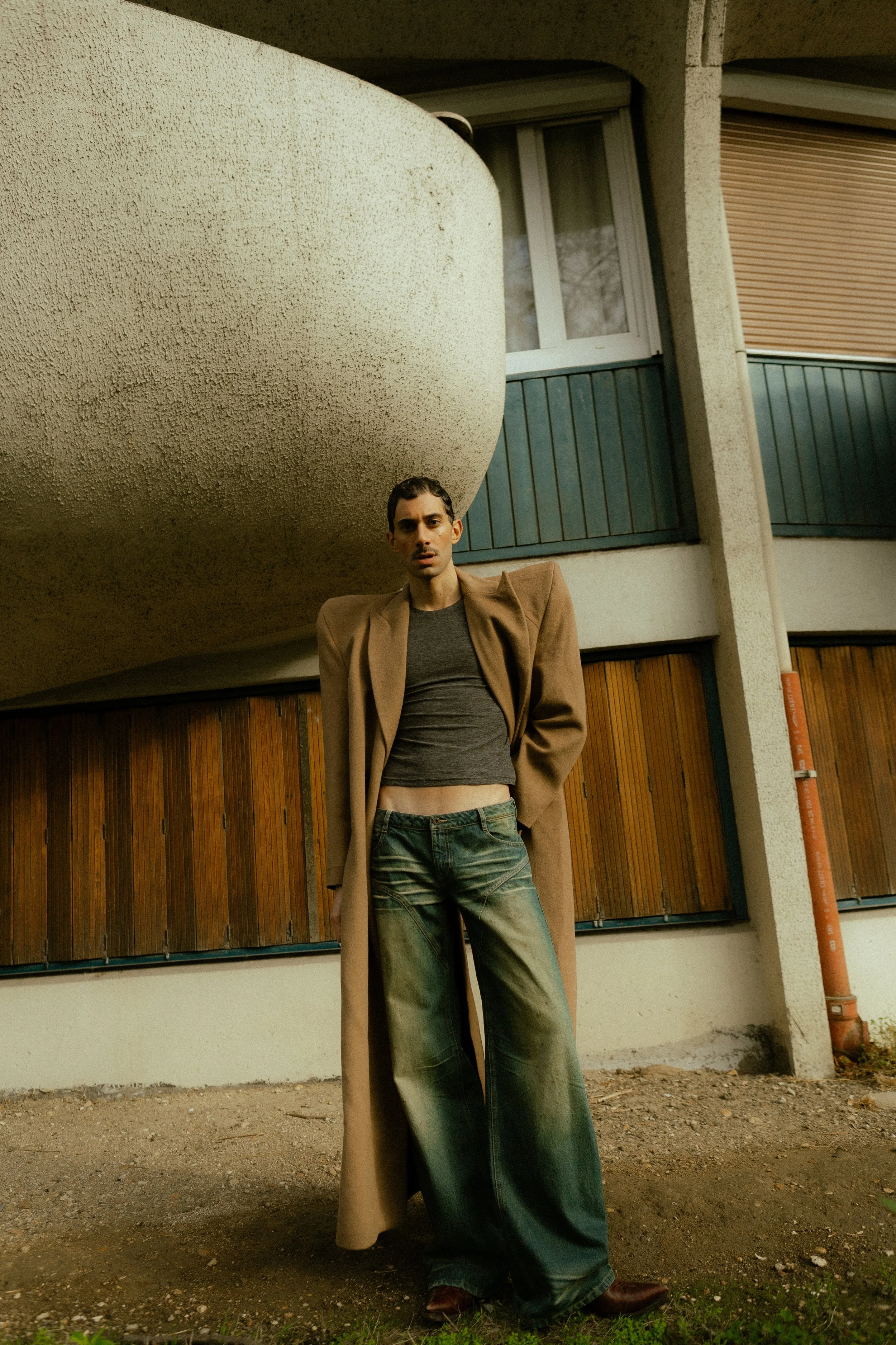Un homme portant un long manteau beige et un pantalon ample se tient devant un bâtiment à l'architecture moderne. Le cadre inclut une fenêtre et un mur texturé.
