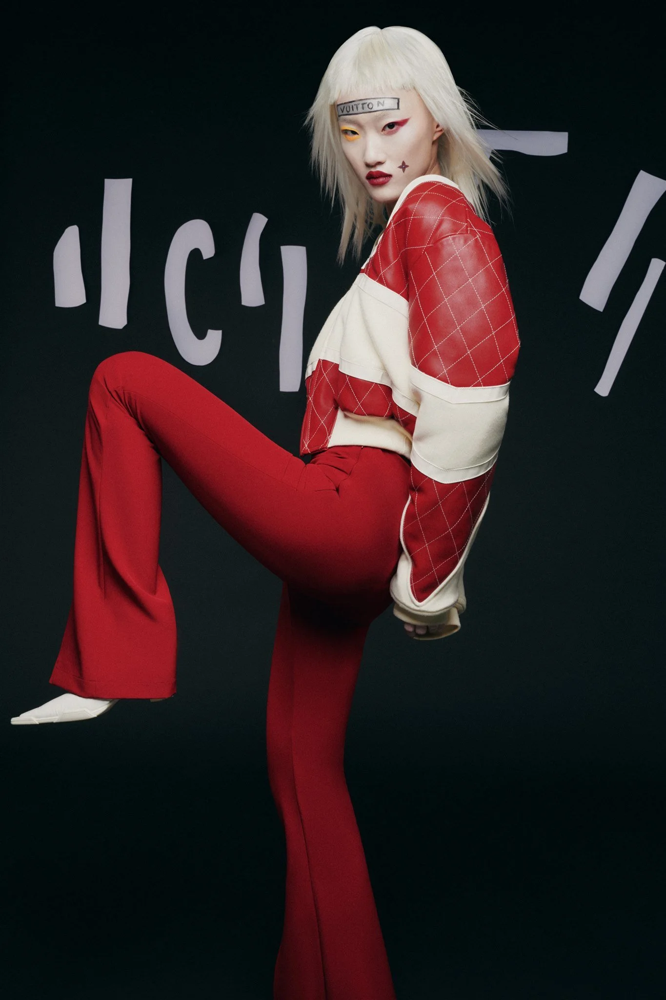Une personne aux cheveux blonds, portant un vêtement rouge et blanc, pose de profil avec une jambe levée devant un fond noir décoré de formes blanches abstraites.