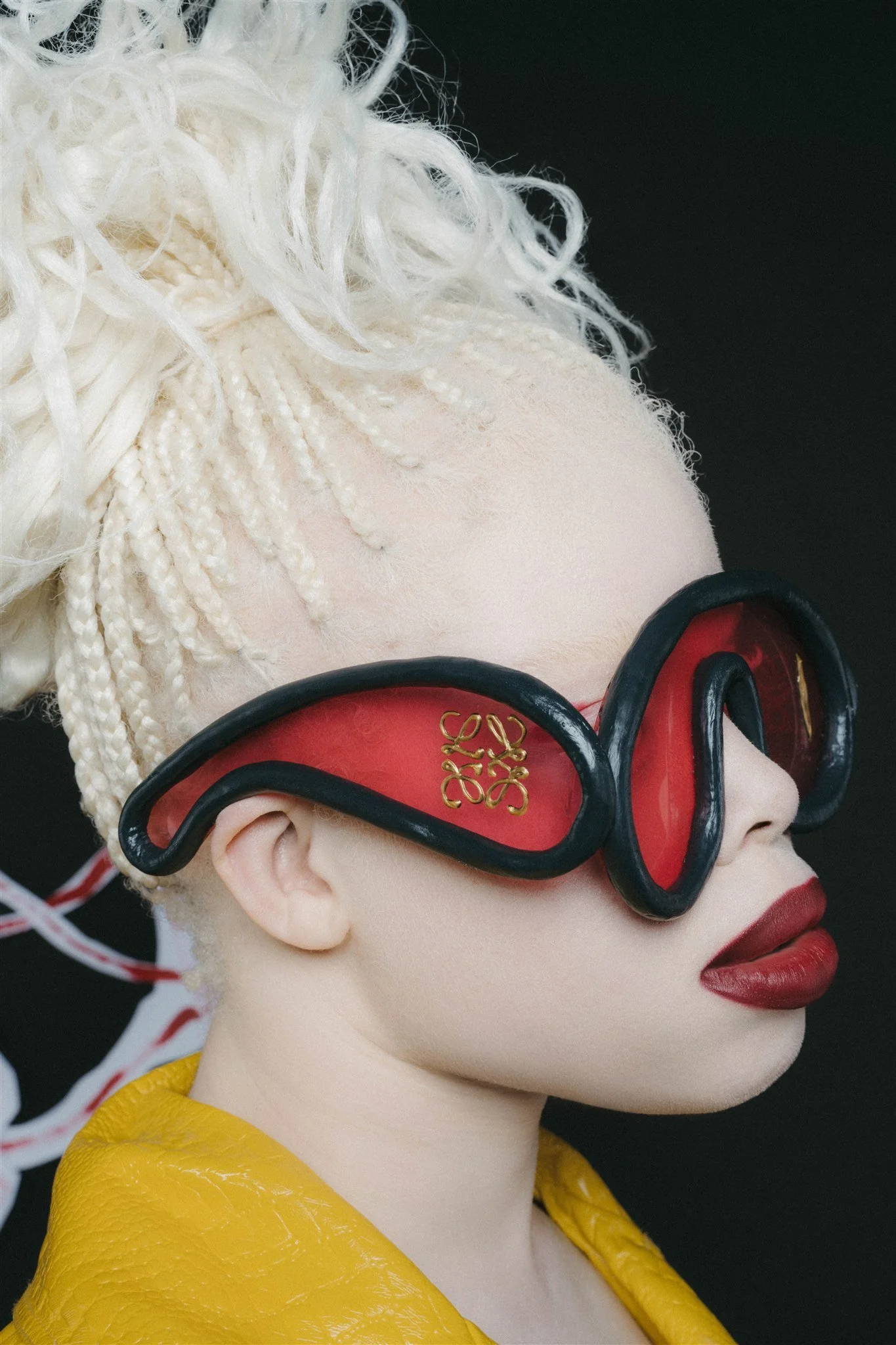 Personne portant des lunettes rouges décoratives et un manteau jaune, avec des cheveux blonds tressés et un rouge à lèvres foncé.