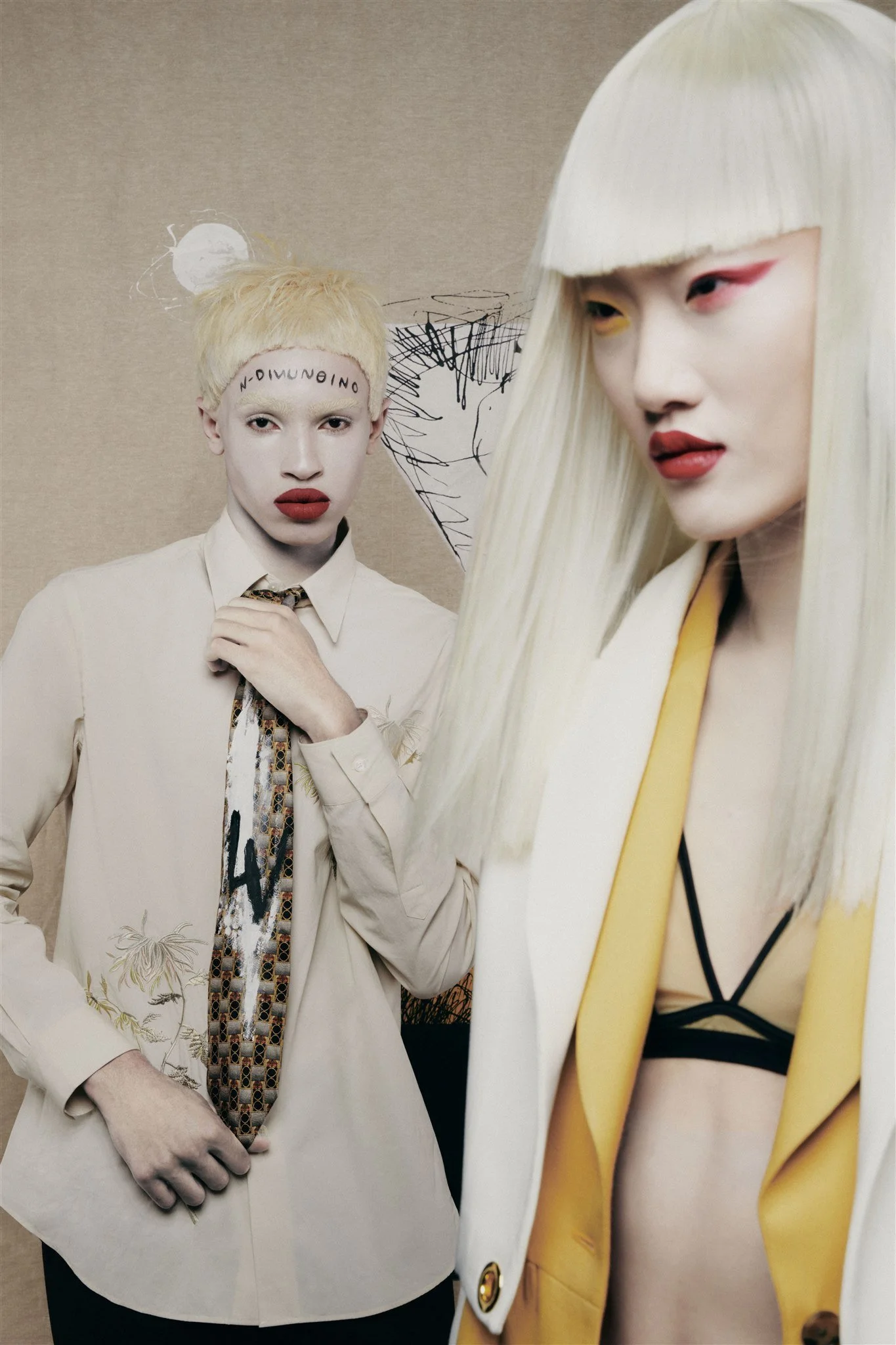 Deux modèles avec un maquillage artistique et des coiffures blondes posent devant un fond avec des dessins abstraits.