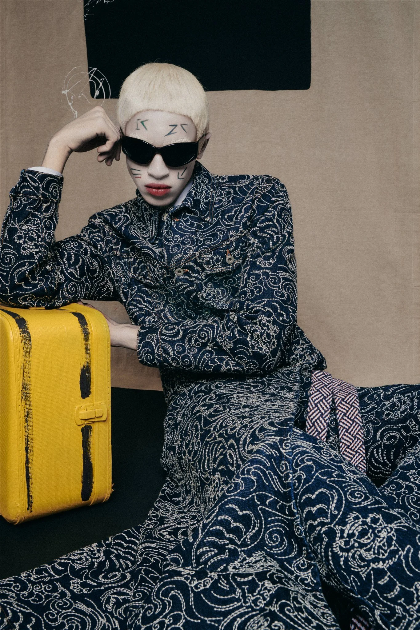 Personne en tenue brodée complexe, lunettes de soleil noires, marquages faciaux; pose avec une valise jaune.