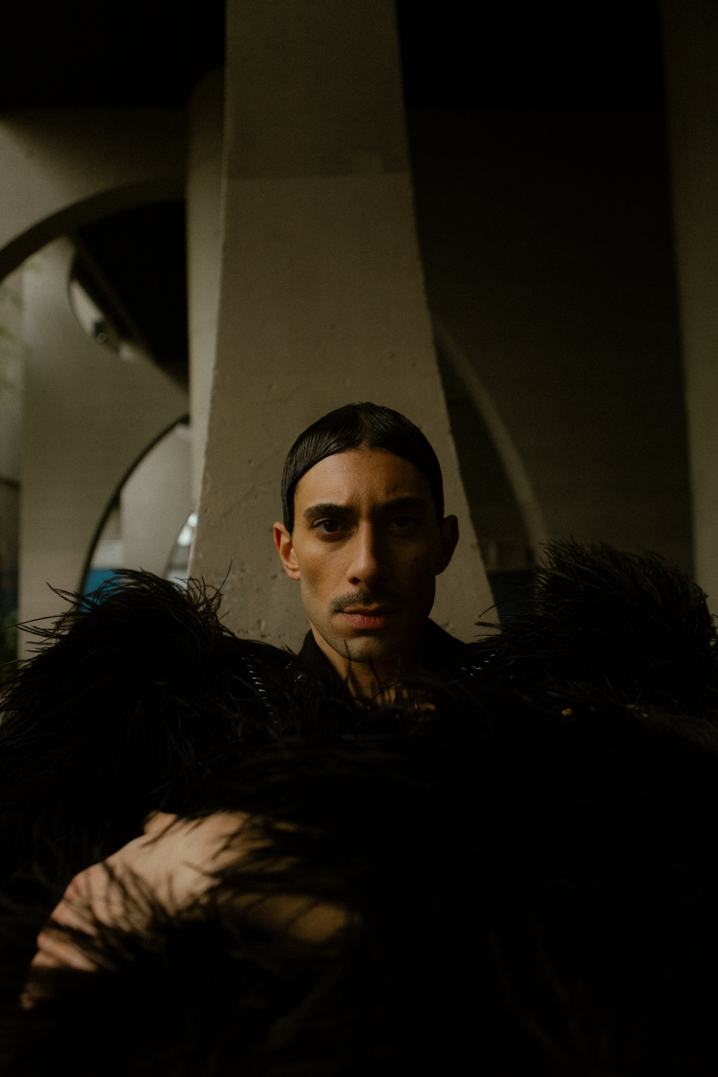 Un homme se tient devant un pilier, portant un vêtement noir à plumes. Il a une expression sérieuse et est dans un lieu intérieur avec une lumière tamisée.