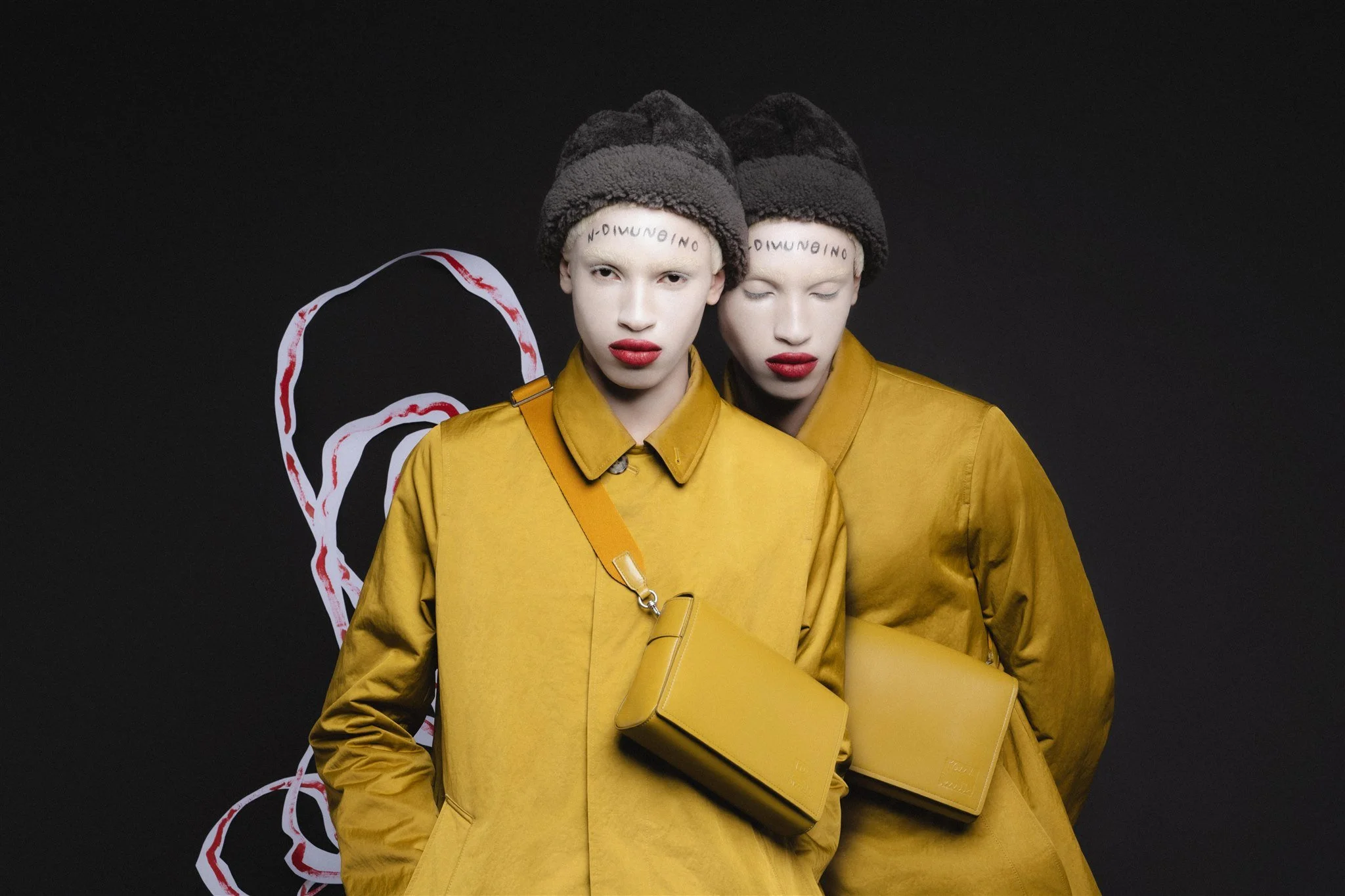 Deux personnes portant des manteaux jaunes, avec des sacs assortis, posant devant un fond noir. Elles ont un maquillage rouge vif sur les lèvres et un texte écrit sur le front.