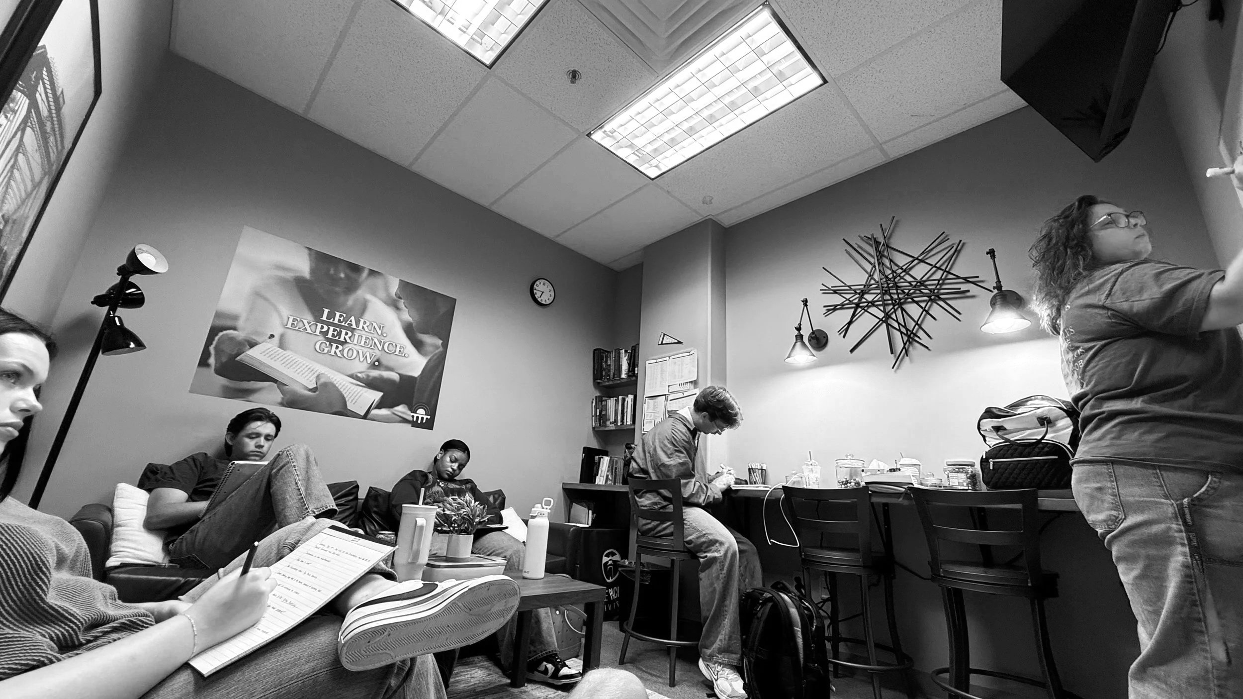A grayscale photo of a group of people in a small room, some sitting on a couch and others at a table. A woman is standing and writing on a whiteboard. The room has a wall clock, a poster with the words 'Learn. Experience. Grow.', and shelves with books. Overhead fluorescent lighting and decorative wall art are visible.
