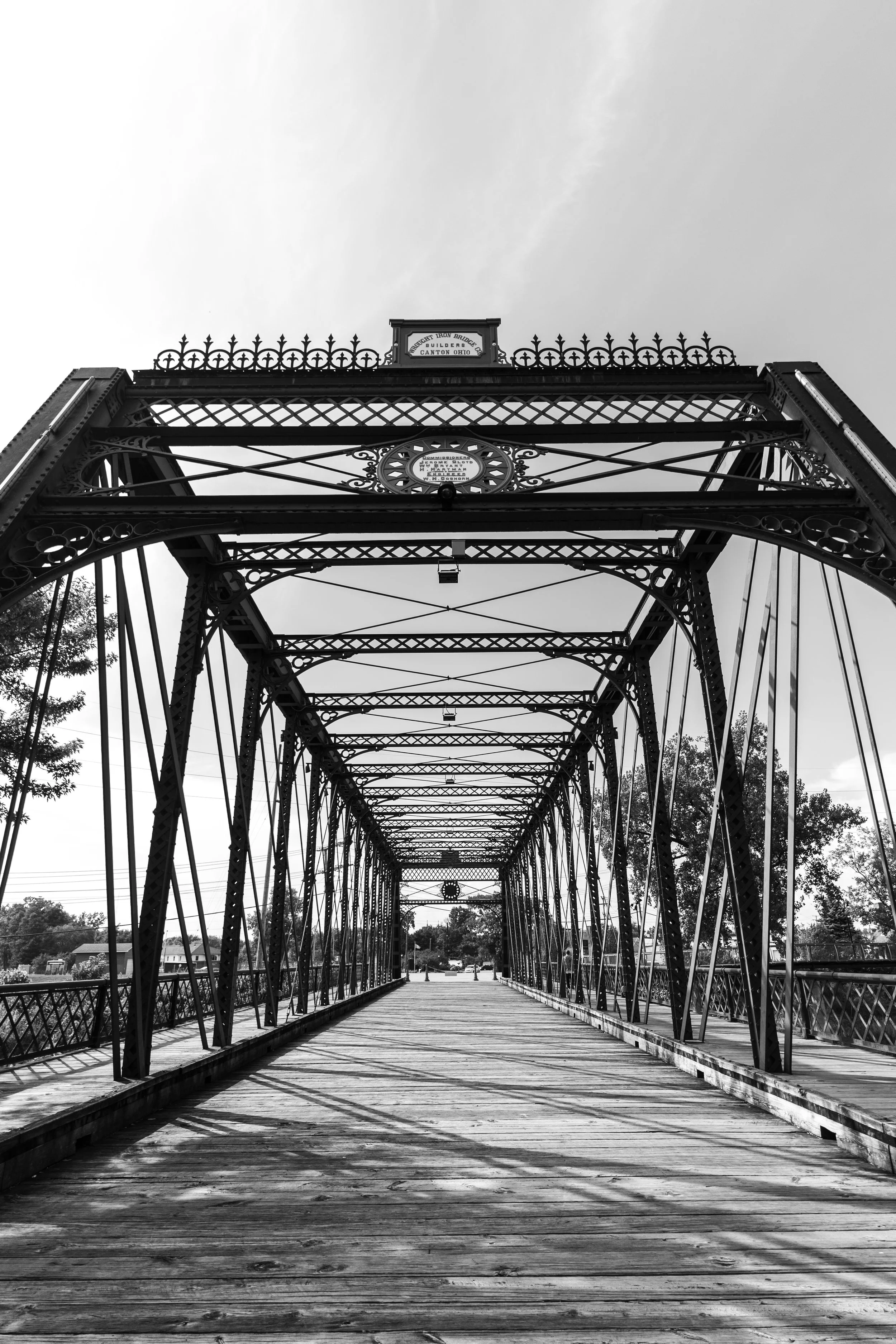 Black and white photo of a historic metal truss bridge crossing a river, with trees and houses in the background.