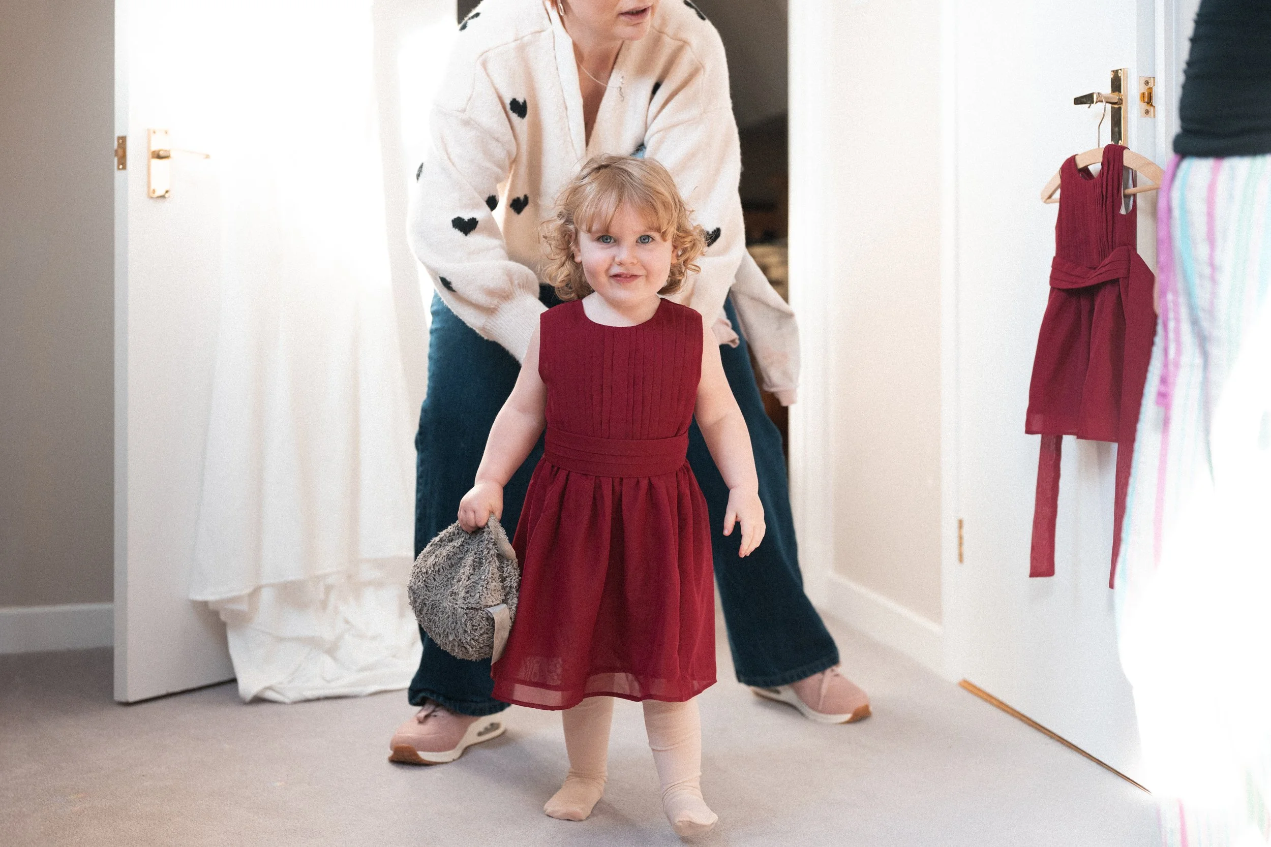 A young girl in a red dress holding a gray stuffed animal, standing in a room with an adult woman behind her, near a door. The girl has curly hair and is smiling.