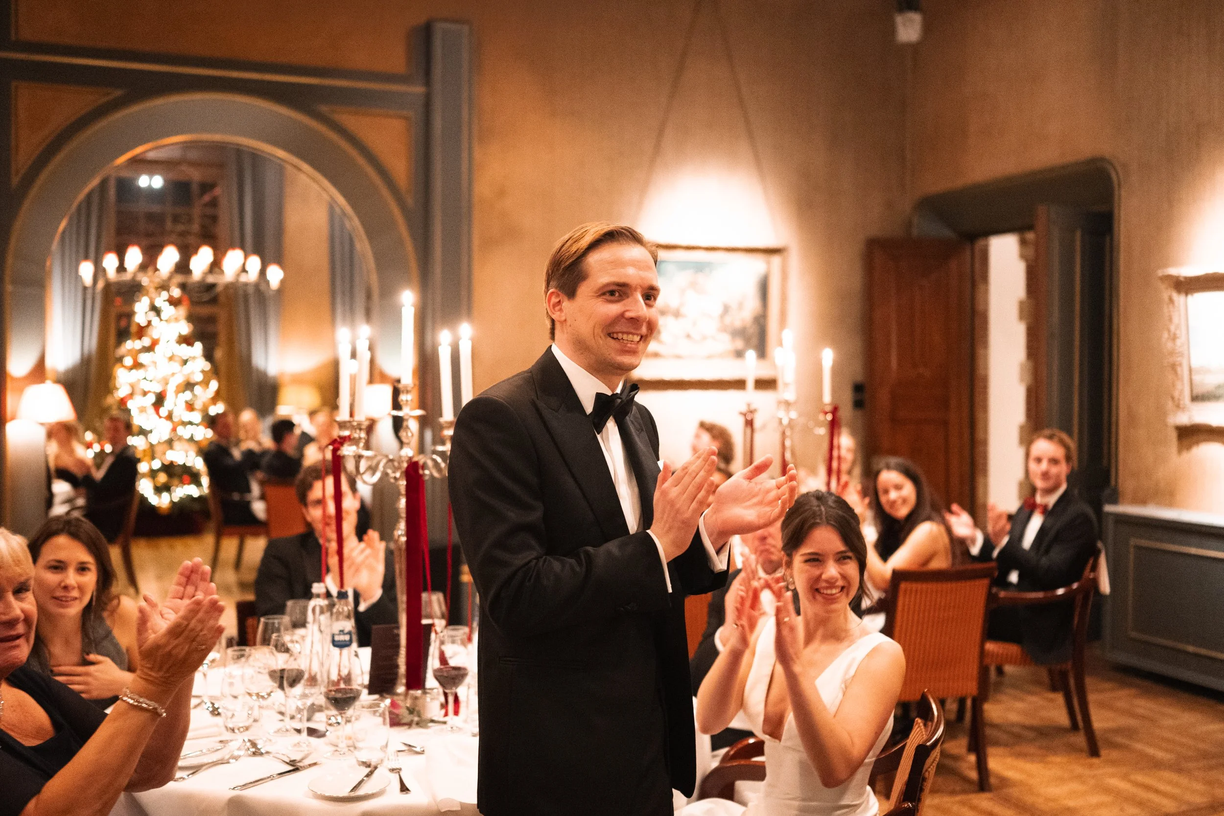 Man in a tuxedo clapping at a wedding reception with seated guests around a decorated table and a Christmas tree in the background.