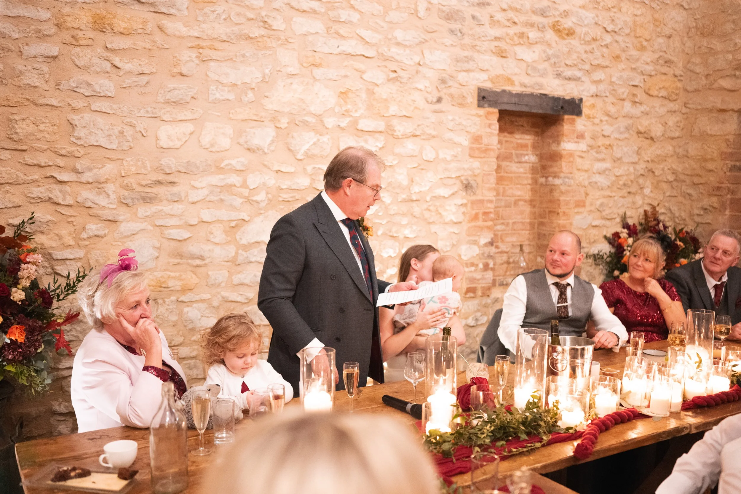 A wedding reception with a man giving a speech at a decorated table with candles and flowers. Several guests, including children, are sitting and listening.