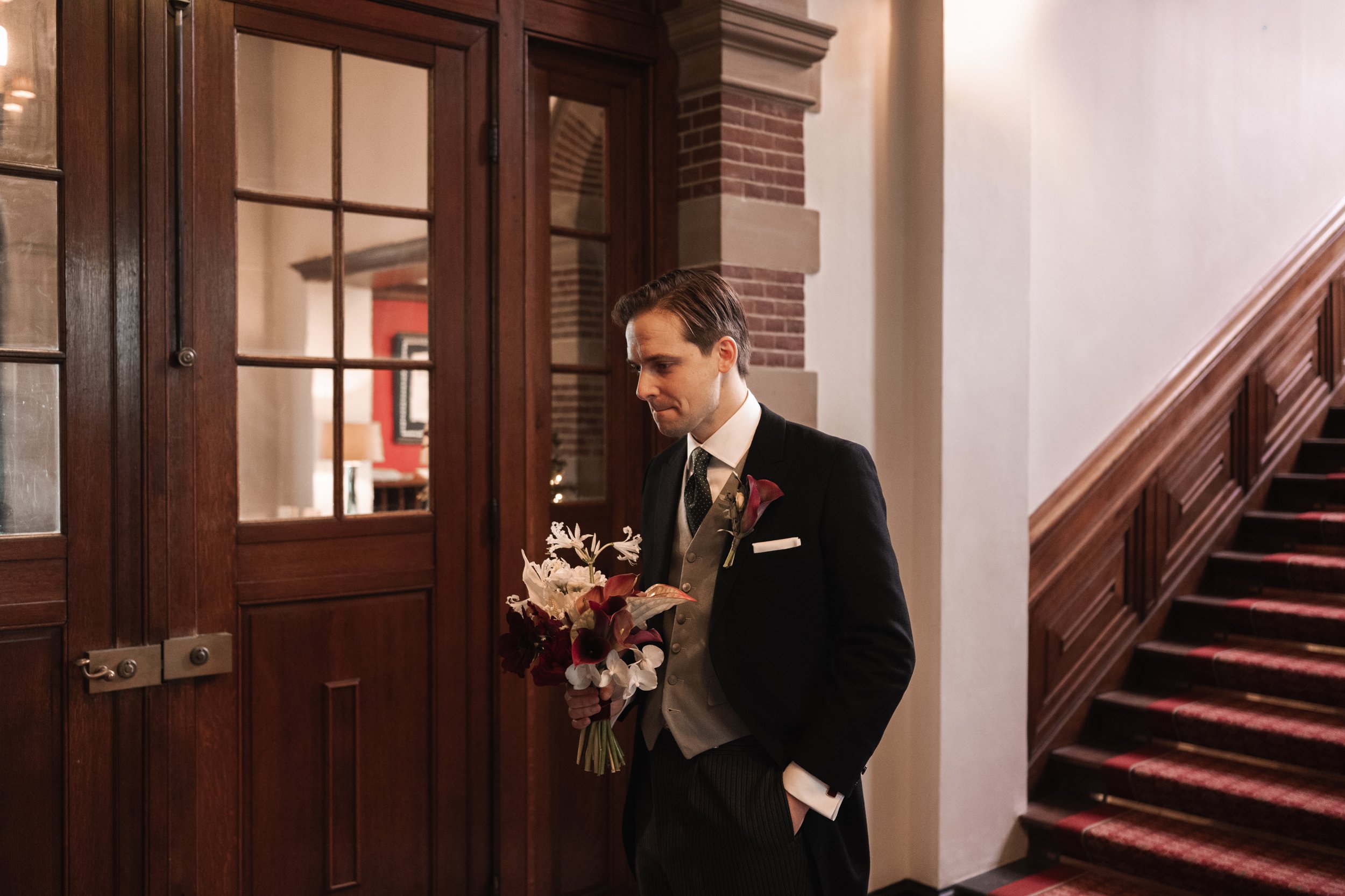 A man in a tuxedo holding a bouquet of flowers indoors near a wooden door and staircase.