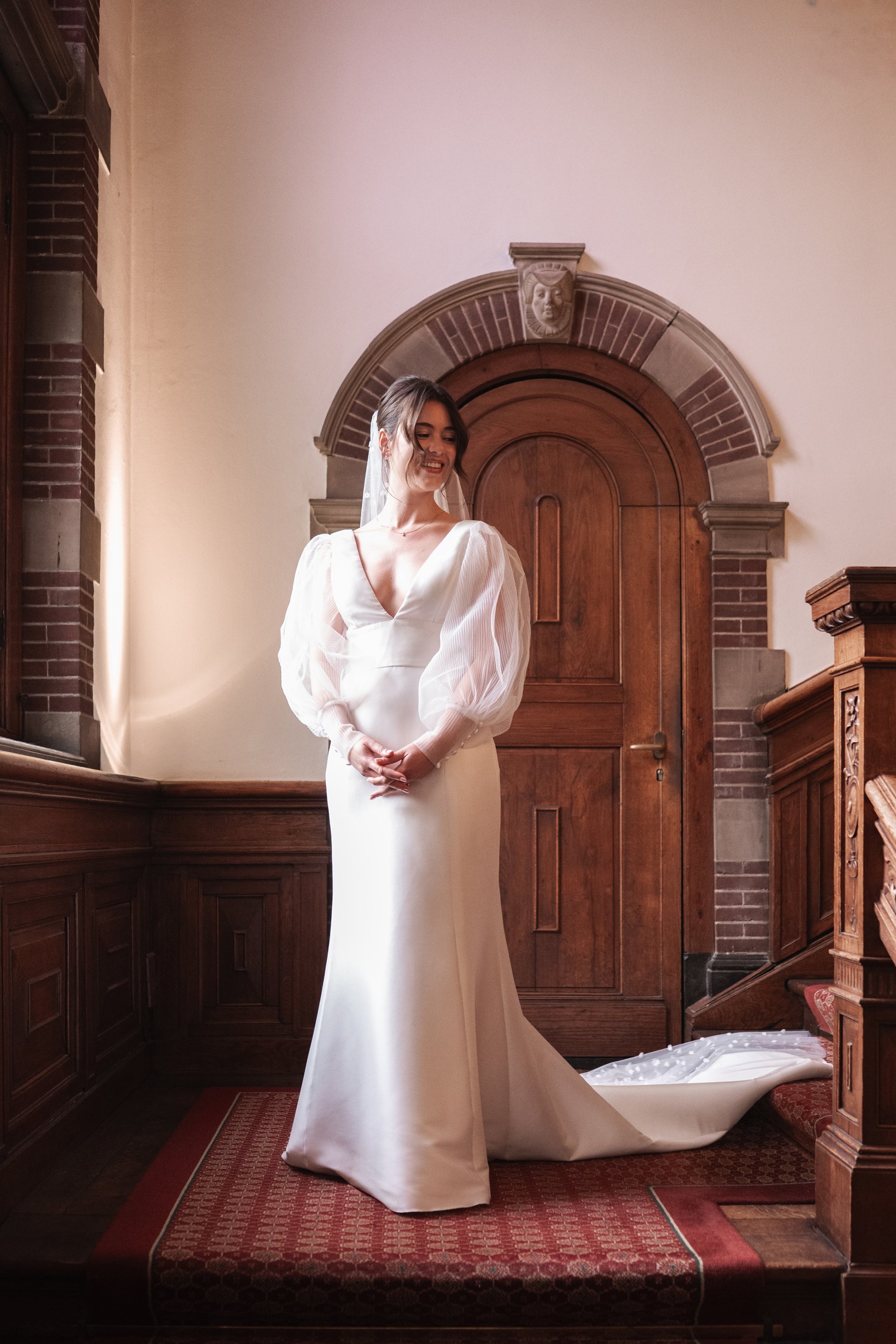 A bride in a white wedding gown with puffed sheer sleeves, standing in front of a wooden door with an ornate brick arch and lion head sculpture, inside a wood-paneled room.