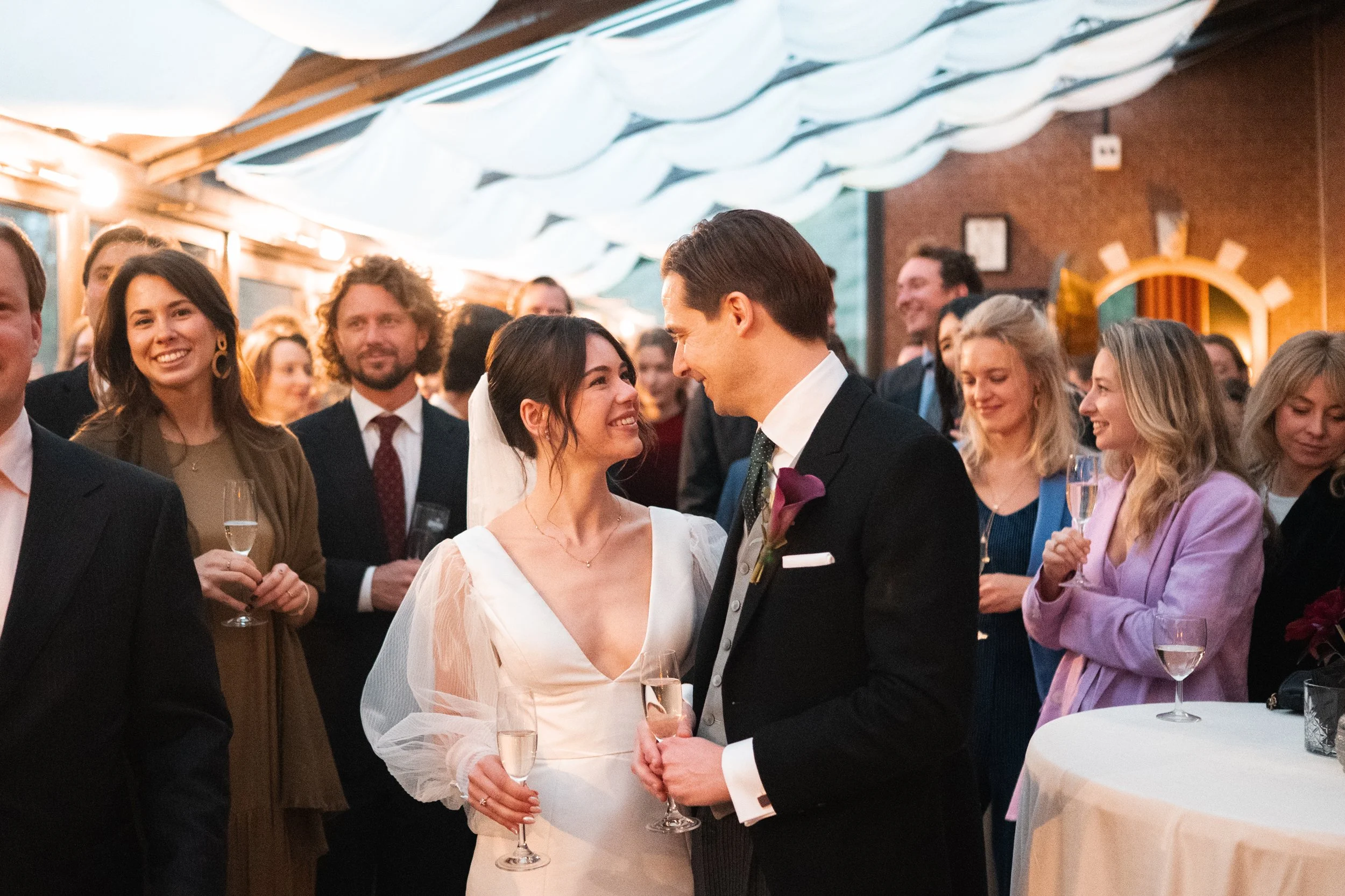 A bride and groom smiling at each other at their wedding reception, surrounded by guests holding champagne glasses.