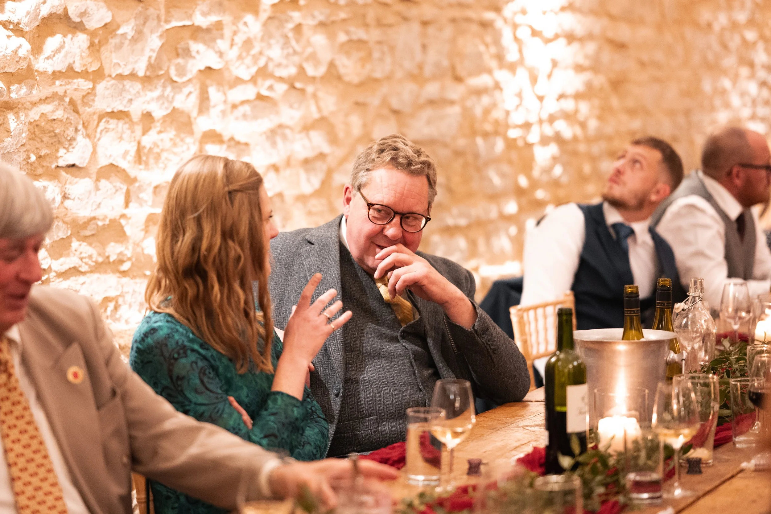 People seated at a dining table with wine bottles, glasses, candles, and a brick wall background, engaging in conversation.