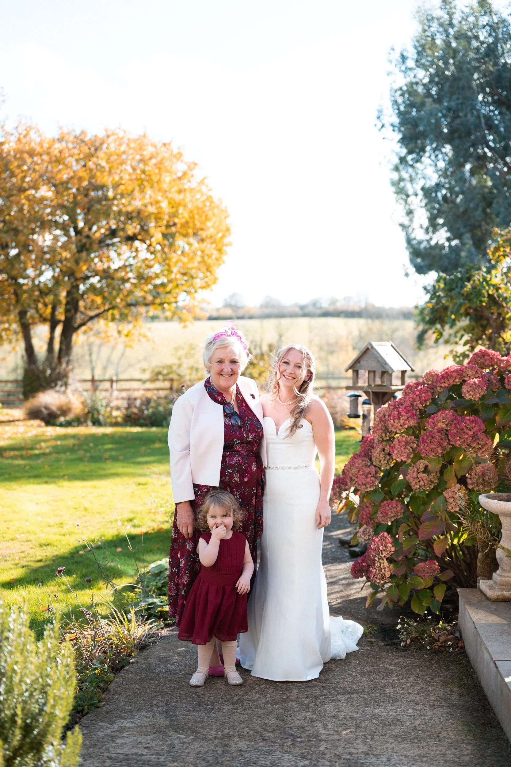 A woman in a white wedding dress and a young girl in a burgundy dress standing with an elderly woman in a floral dress, outdoors on a sunny day with trees and flowers in the background.