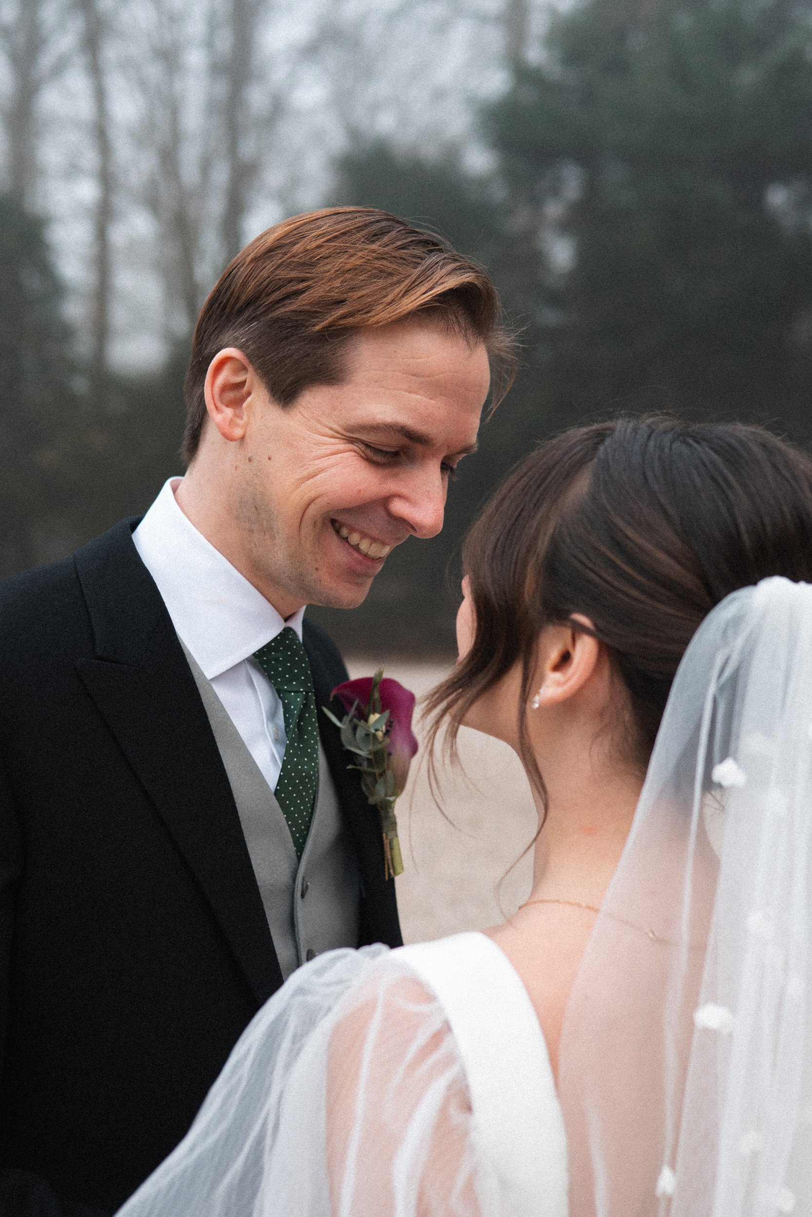 A bride and groom smiling and leaning close to each other outdoors, with trees in the background, during a wedding ceremony.