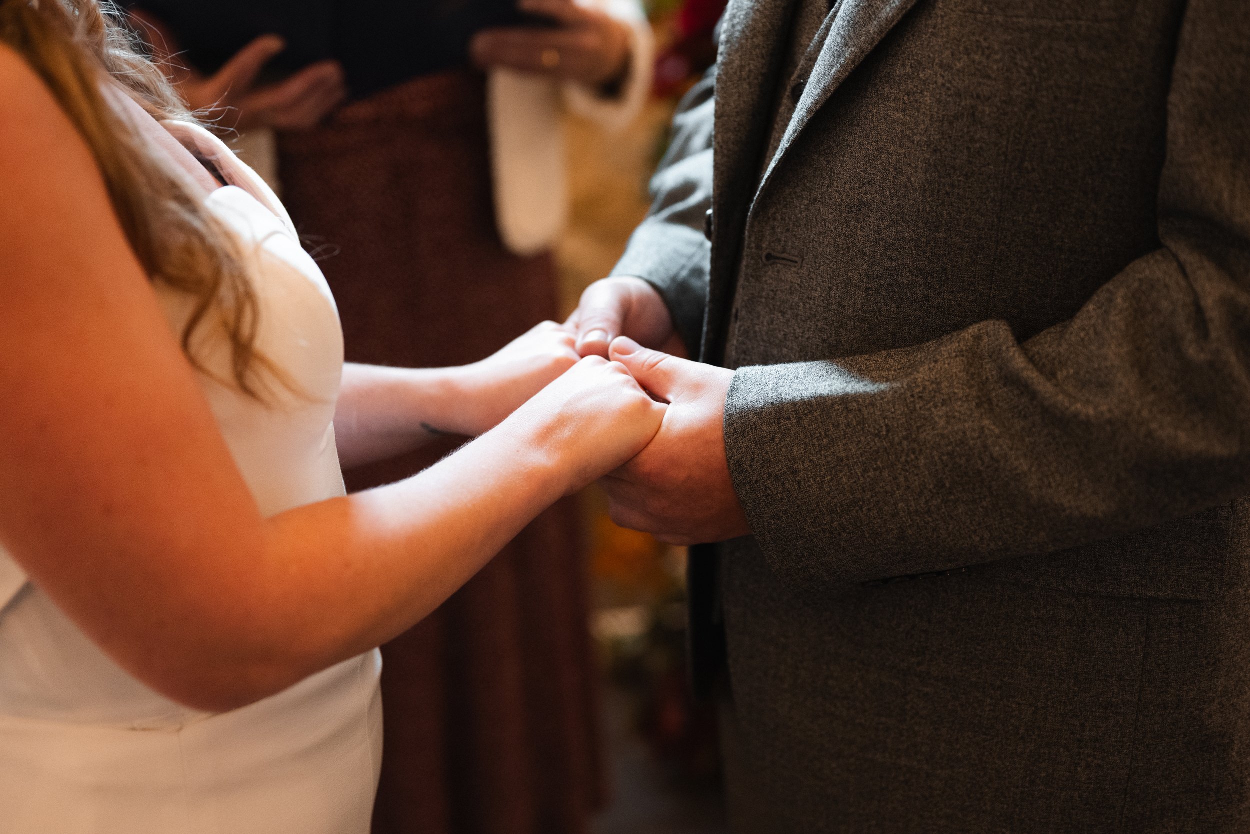A couple holding hands during a wedding ceremony, with the bride in a white dress and the groom in a gray suit.