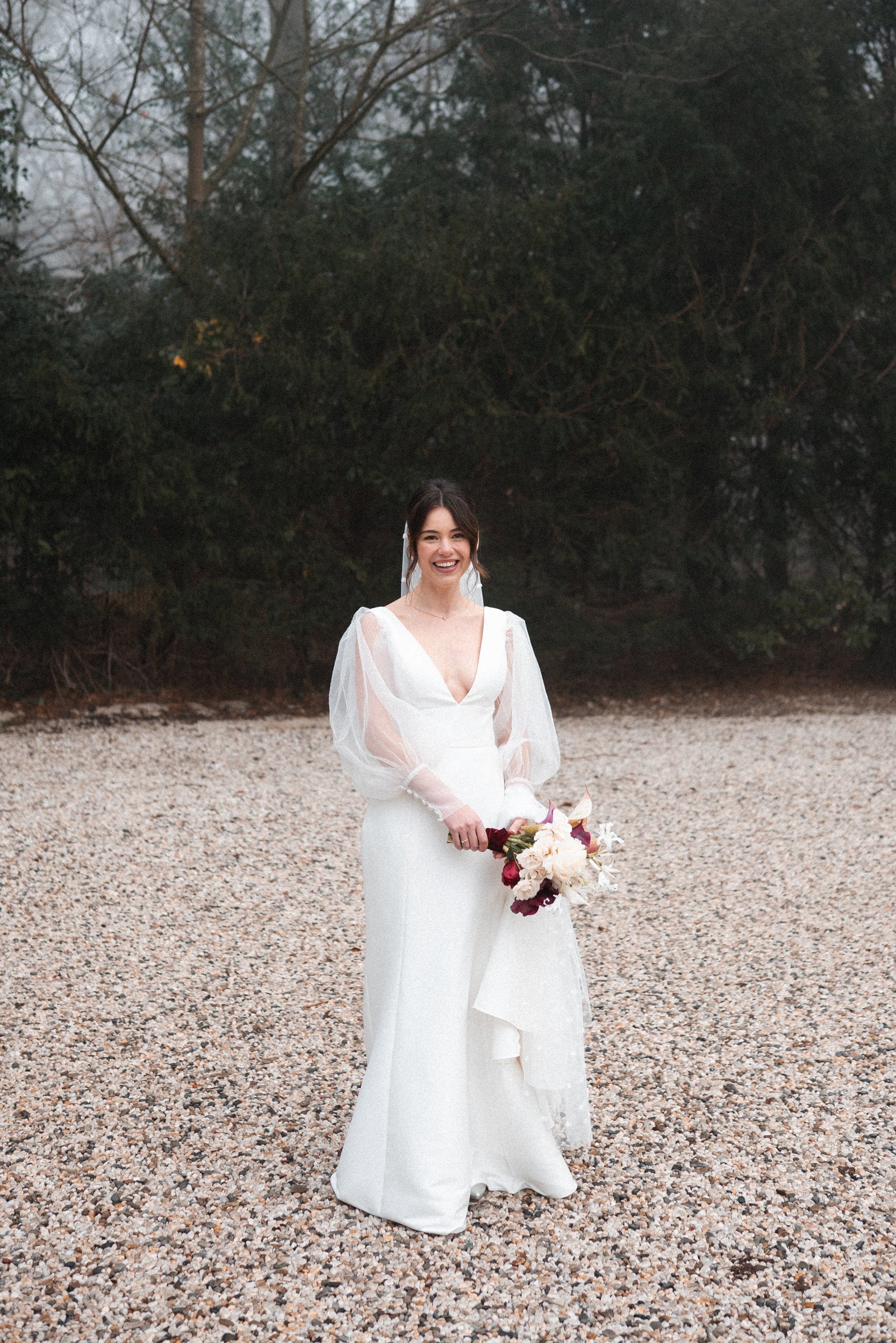 A woman in a white wedding dress holding a bouquet of flowers, standing on a gravel surface outdoors with trees in the background.
