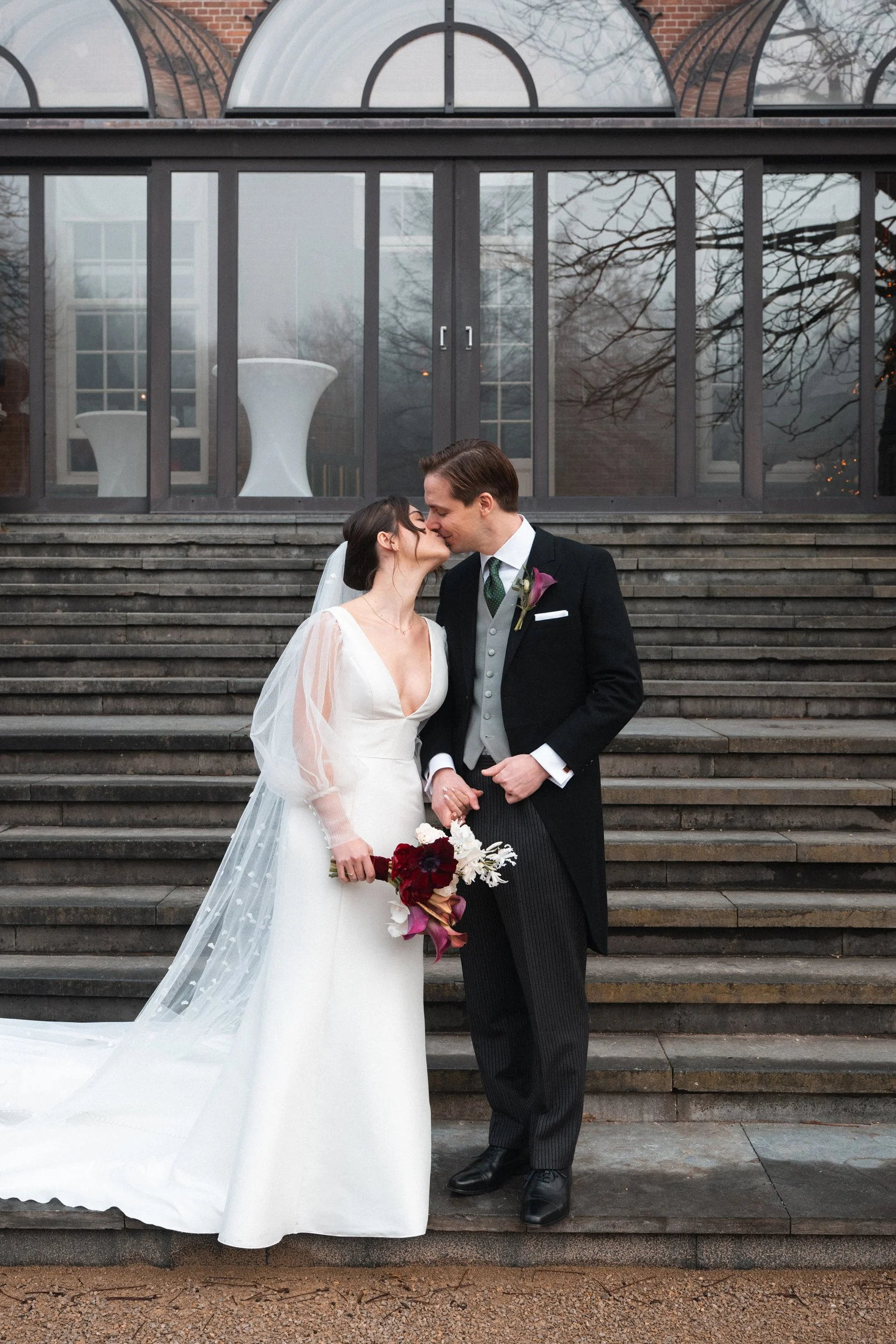 A bride and groom sharing a kiss outside on steps in front of a glass building during their wedding.