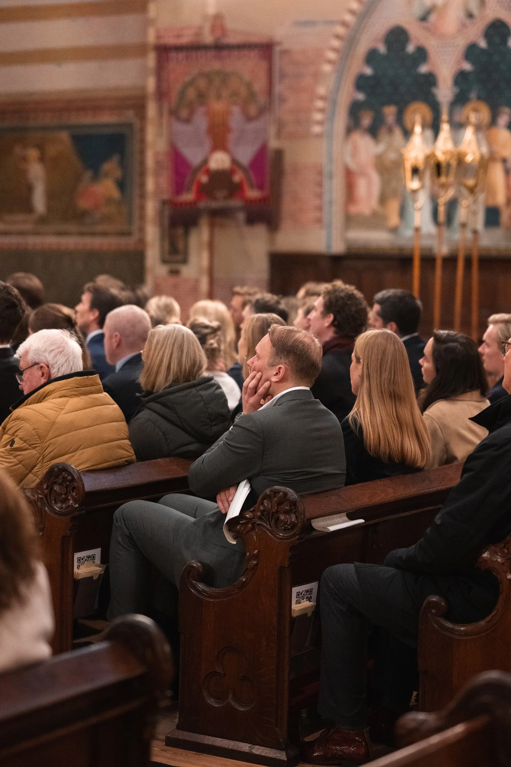 People seated in wooden pews inside a church, attending a service or event, with religious artwork and chandeliers visible in the background.