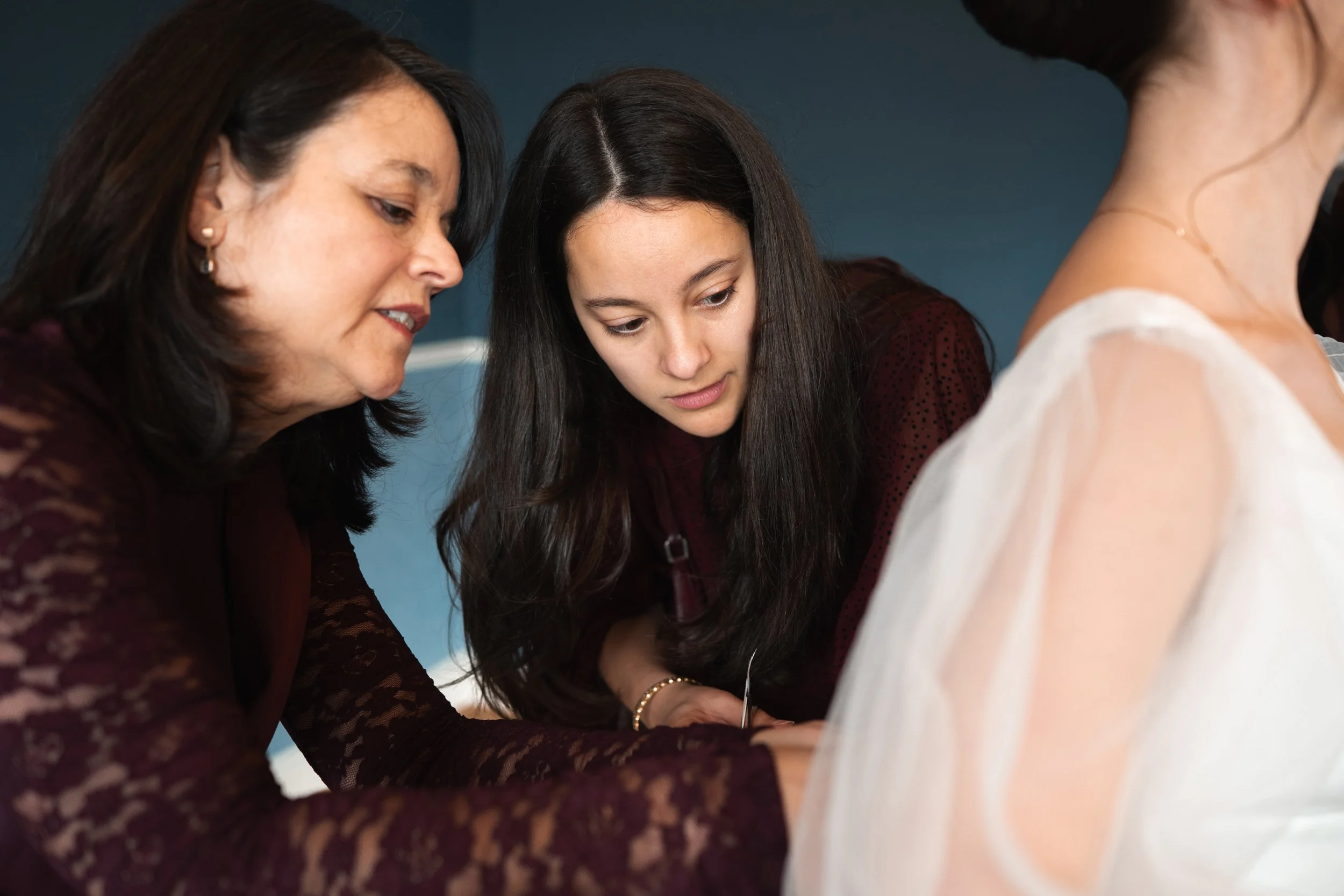 Three women working together closely, focused on an event dress, with a dark blue wall in the background.