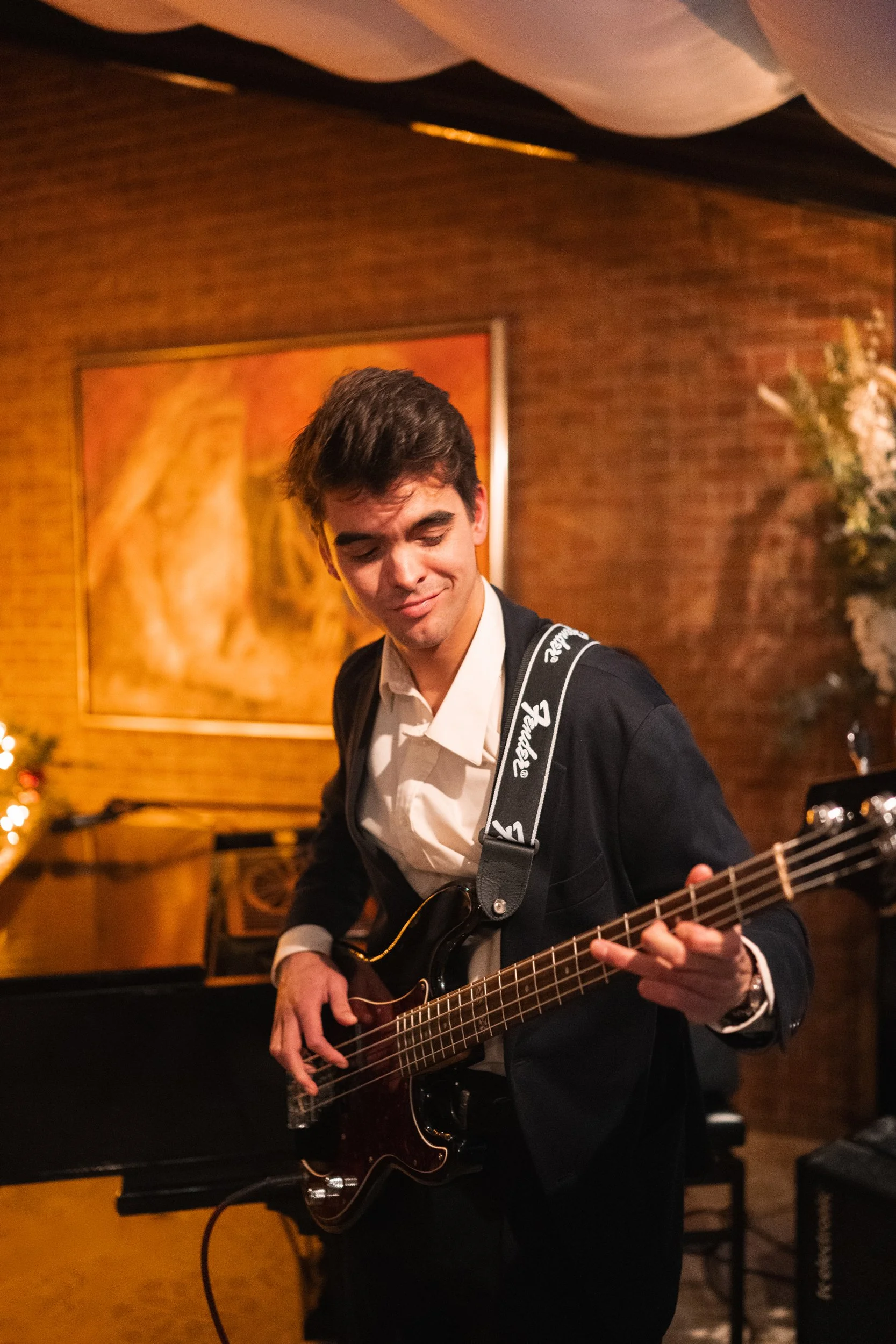 A young man playing an electric bass guitar in a warmly lit room with brick walls and artwork in the background.