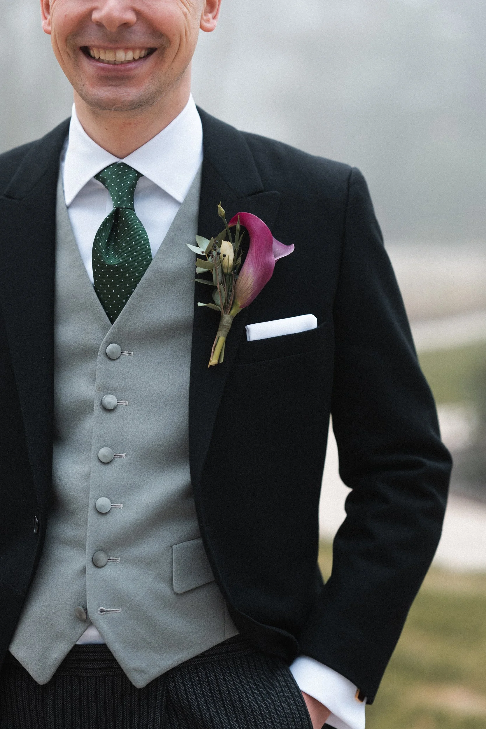Close-up of a smiling man in formal wedding attire, wearing a black tuxedo with a gray vest, white shirt, and green polka dot tie. He has a boutonniere with pink and purple flowers on his left lapel and a white pocket square.