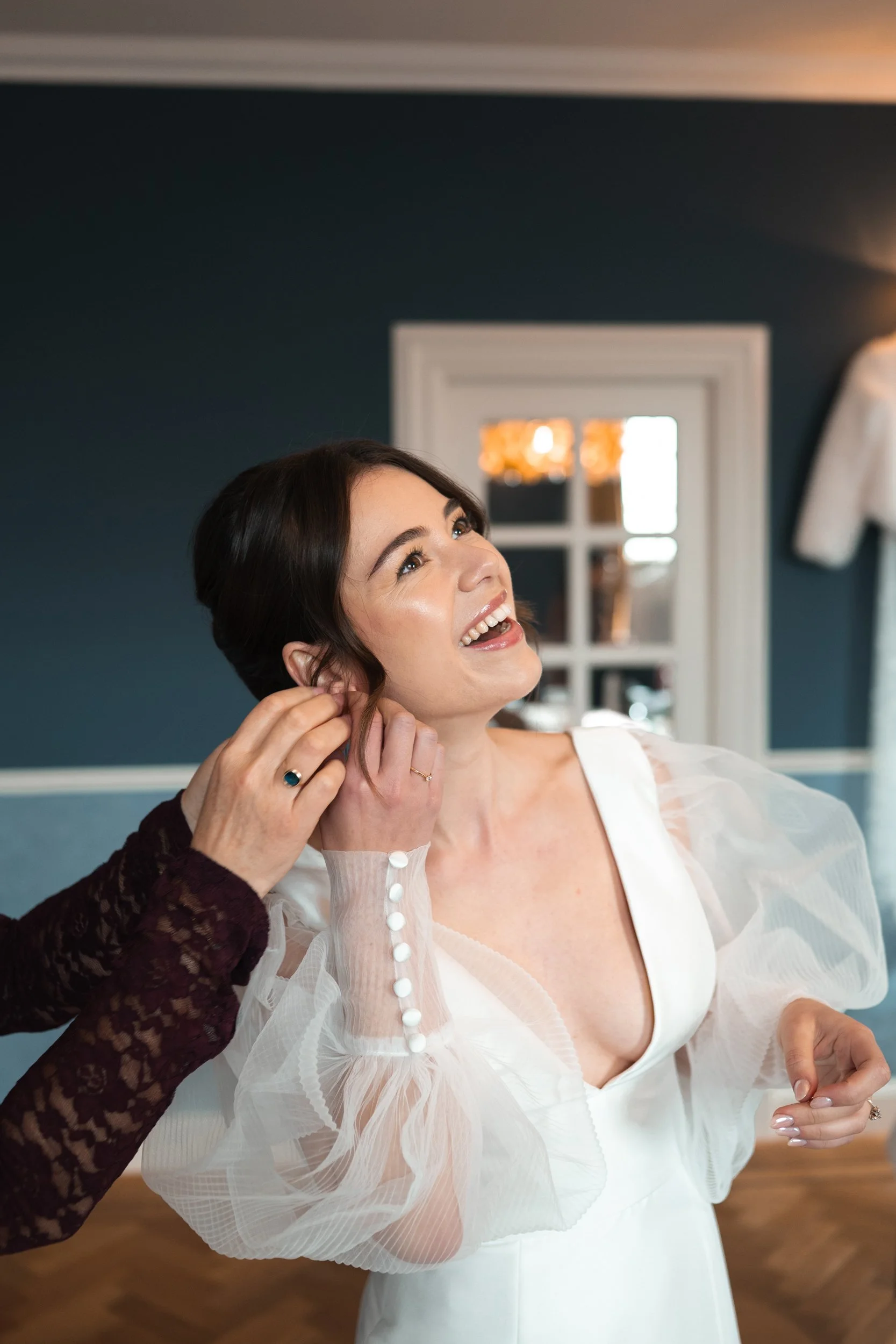 A woman in a white dress is getting ready, smiling while someone adjusts her earring in a room with dark blue walls and a decorative mirror in the background.