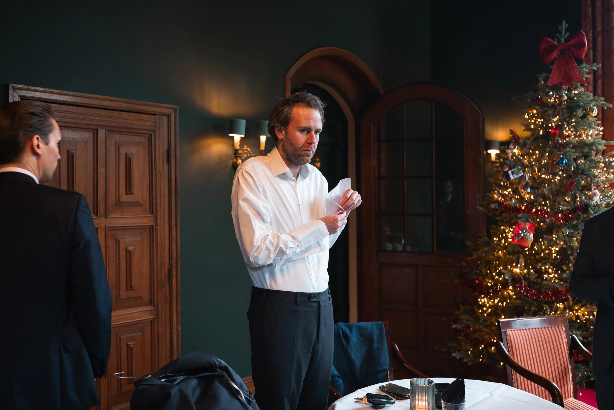 A man in a white shirt reads a piece of paper at a Christmas party, with a decorated Christmas tree in the background.
