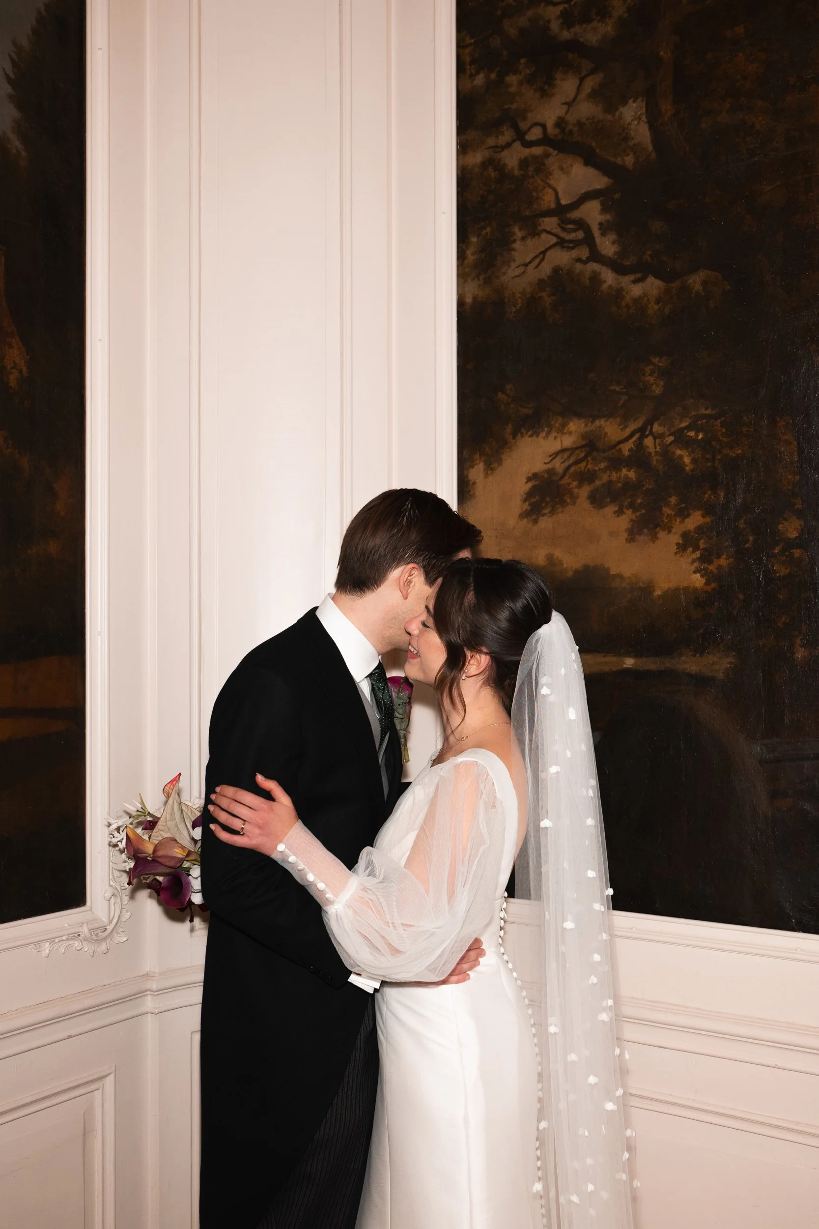 A bride and groom share an intimate moment indoors, standing close with foreheads touching, in front of a wall with dark landscape paintings.