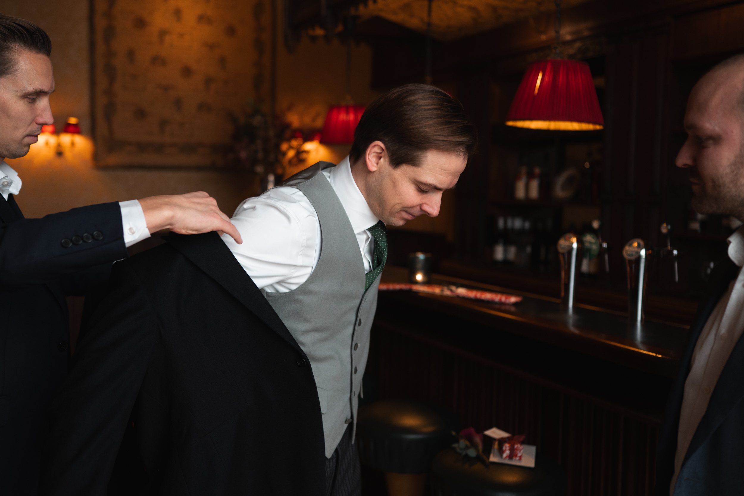 A man in a gray vest and white shirt is being helped to put on a black suit jacket by another man in a black suit at a bar or pub.