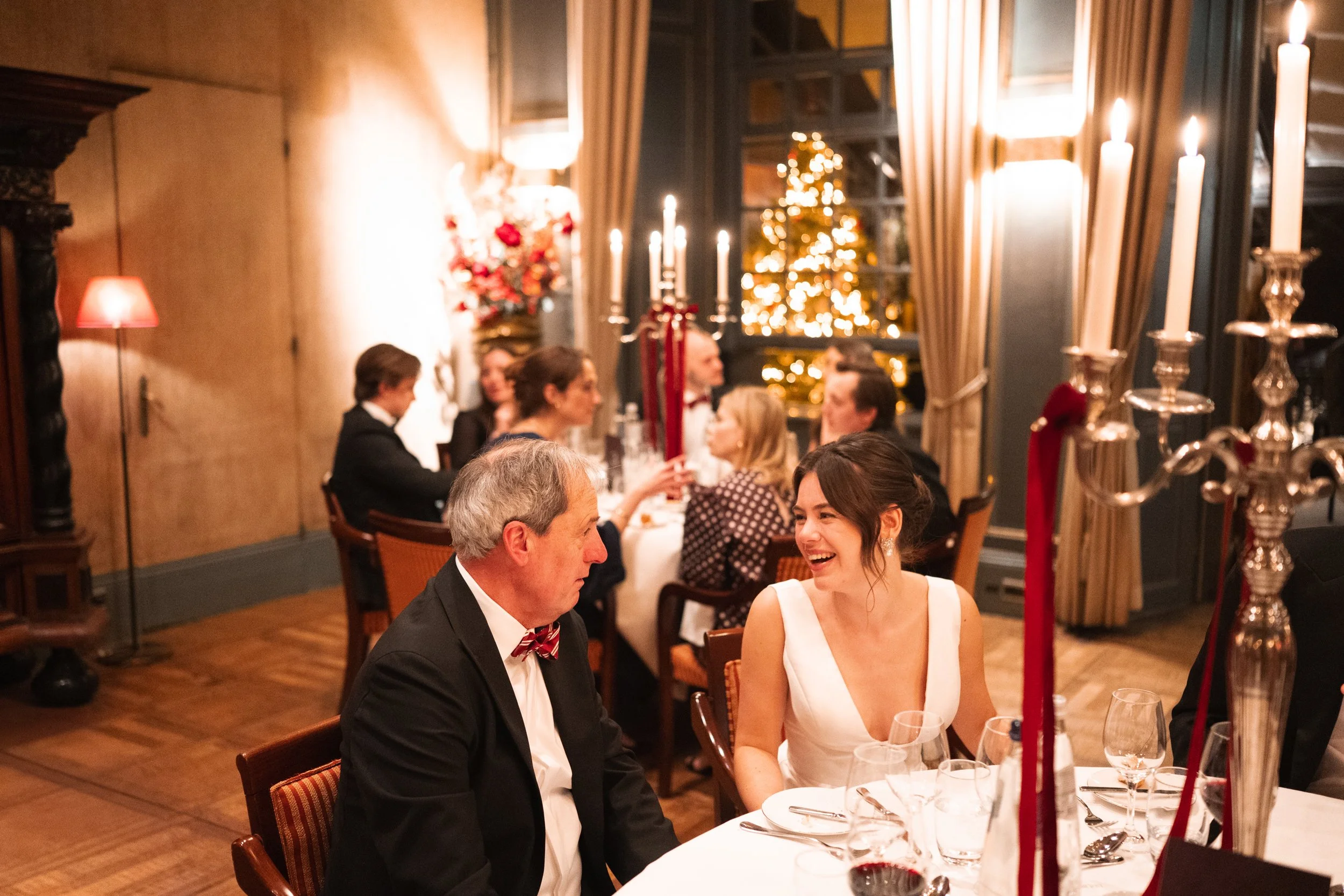 People dressed in formal attire at a Christmas dinner table with candles and a decorated Christmas tree in the background.