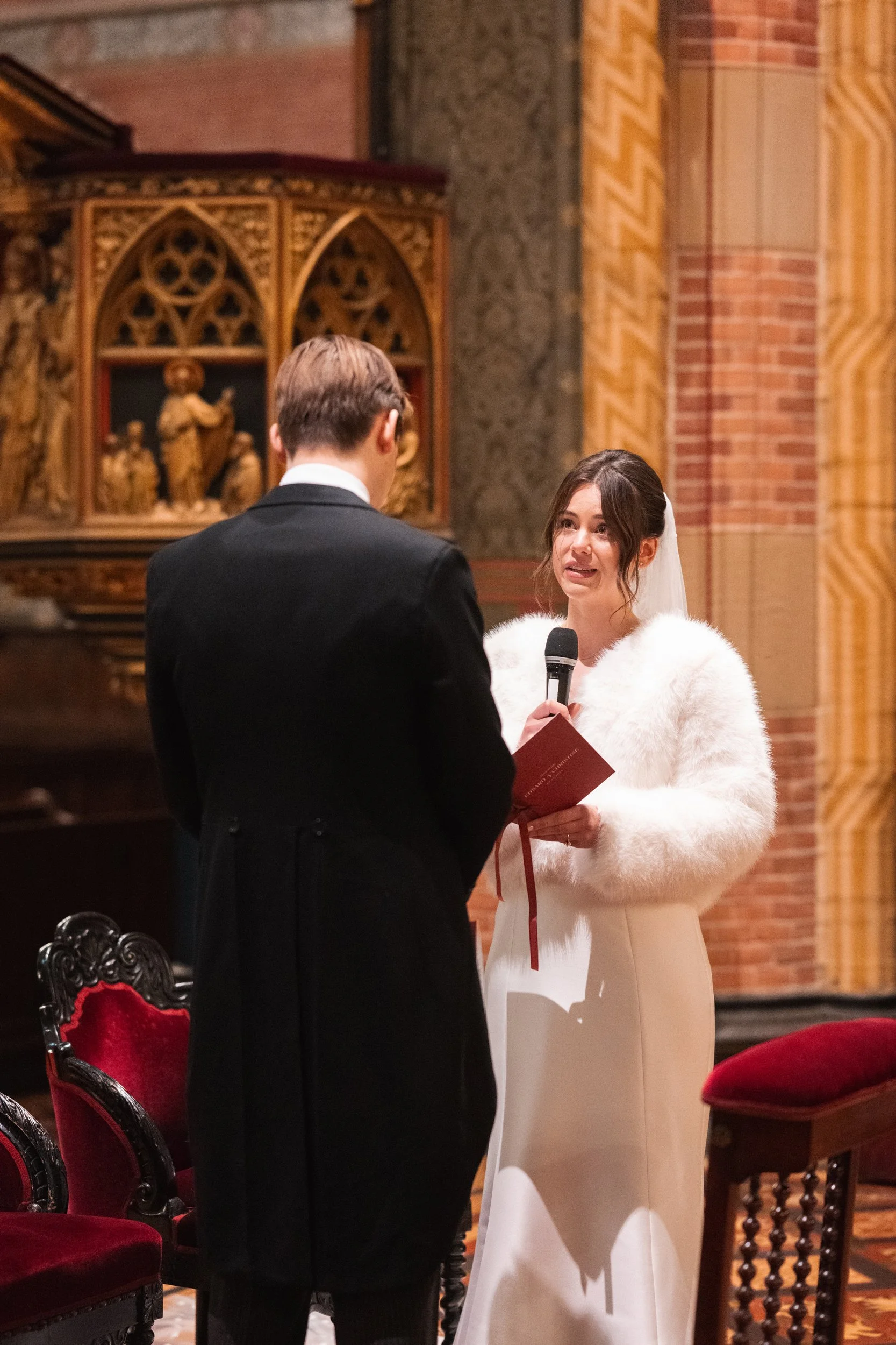 A woman in a white fur jacket and white dress is speaking into a microphone during a wedding ceremony, while a man in a black suit with a white shirt faces her. They are inside a church with ornate wooden decorations and red velvet chairs.